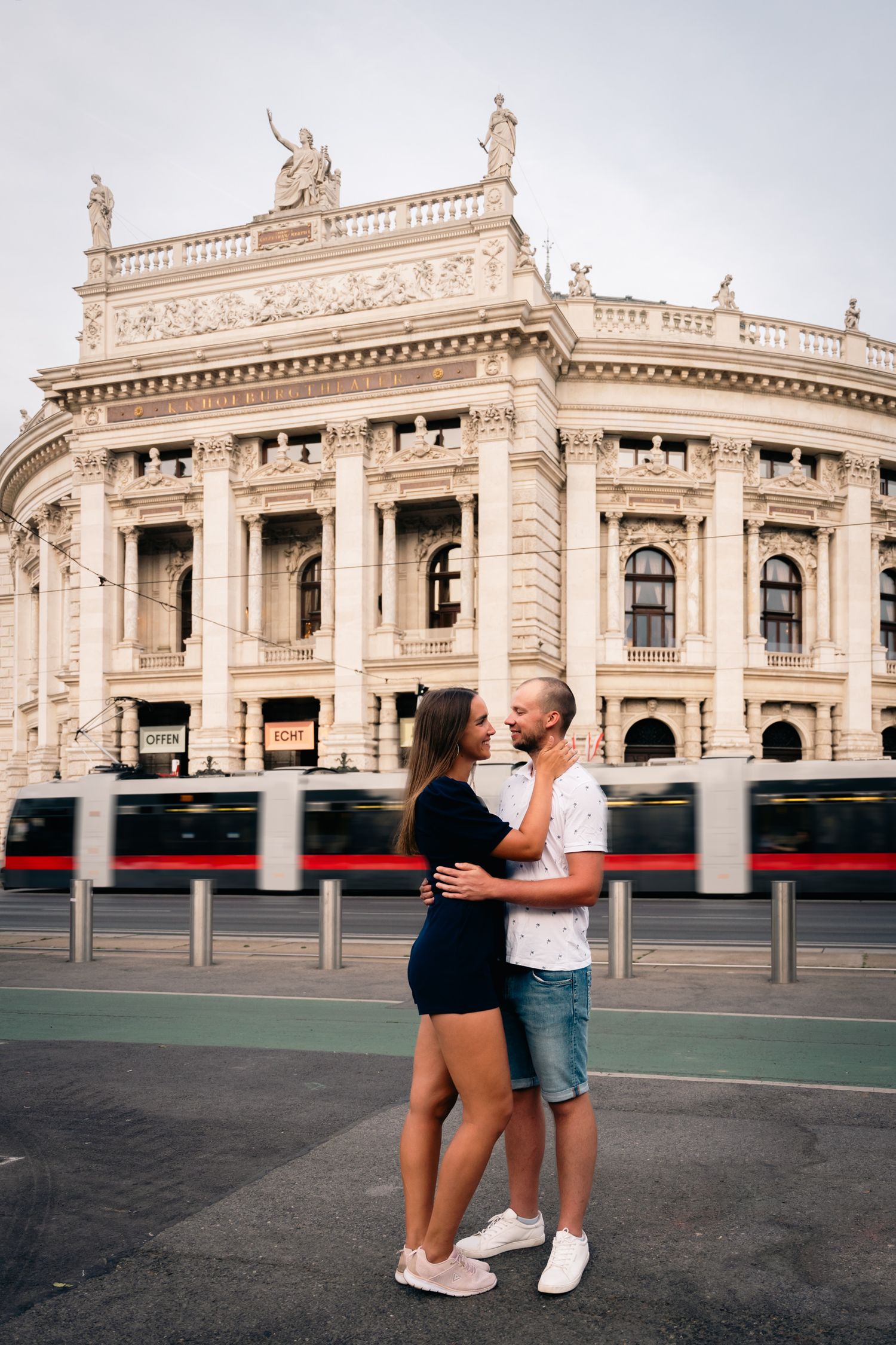 Couple embracing in front of the grand Burgtheater in Vienna with a red tram passing by.