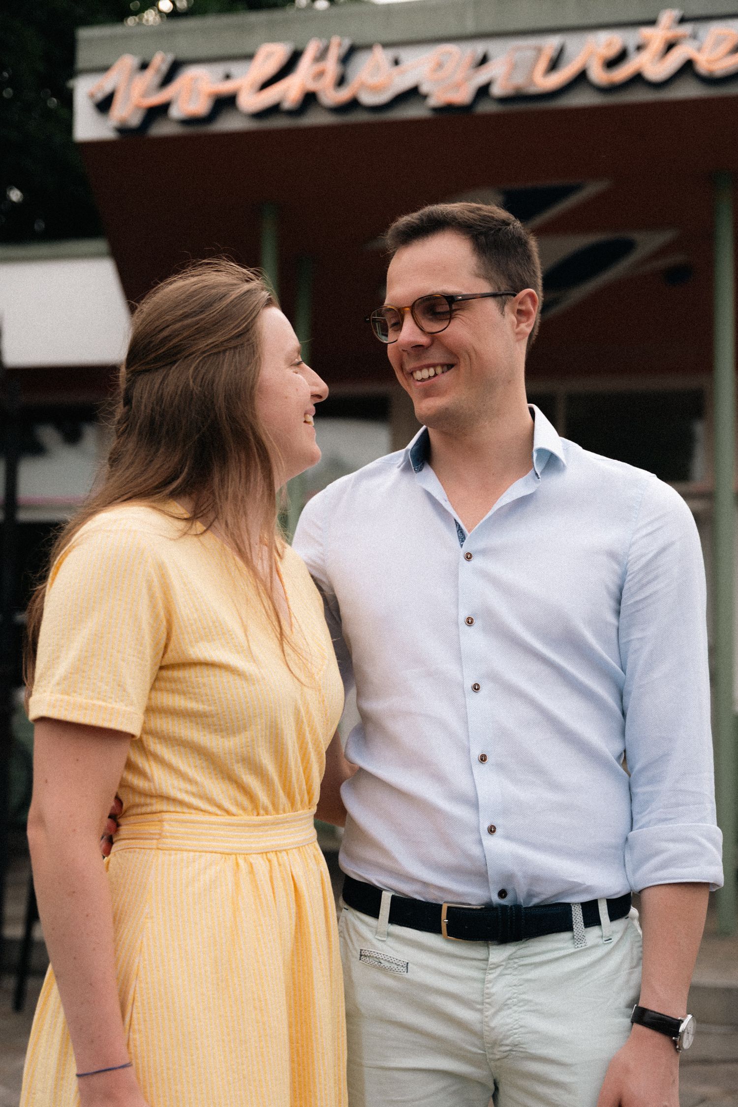 Smiling couple looking at each other outside a retro-style venue with neon signage.