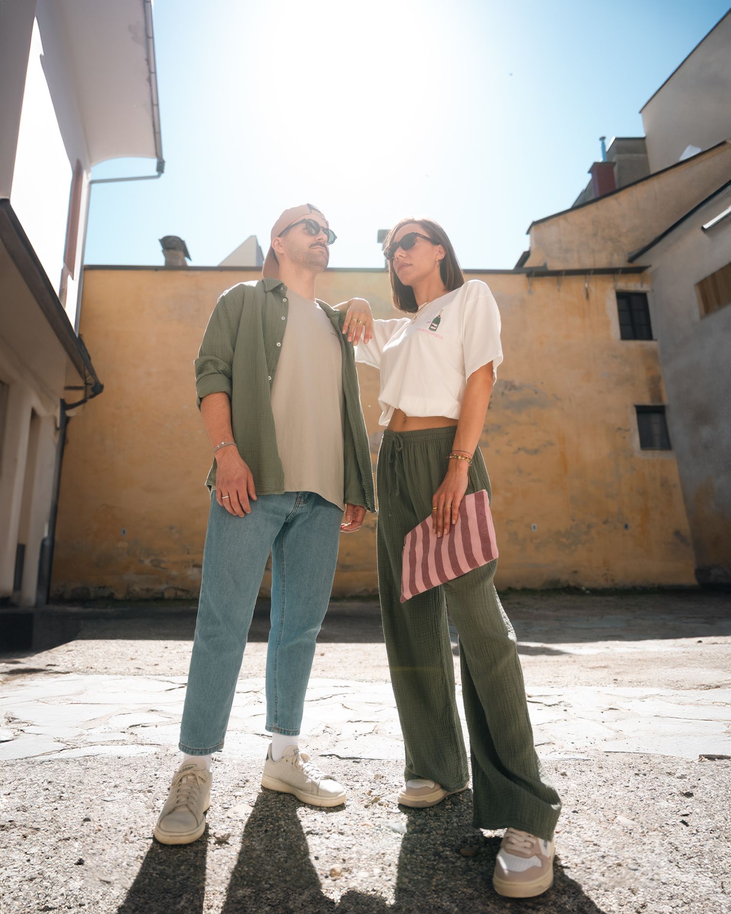 A fashionable couple in casual green and white outfits stand in a sunlit courtyard looking upward.