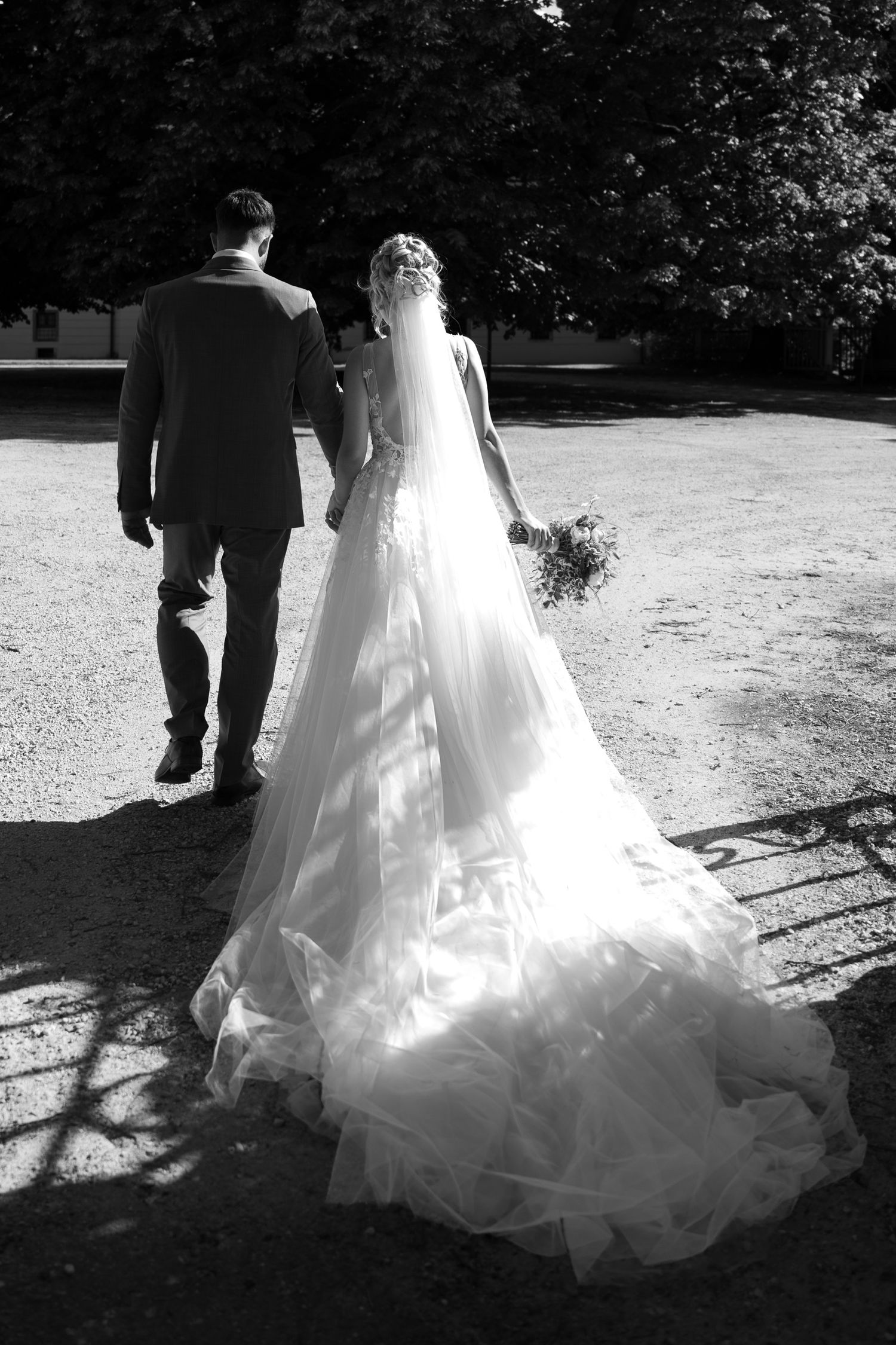 Black and white rear view of bride in flowing gown with long veil walking with groom through sunlit grounds.