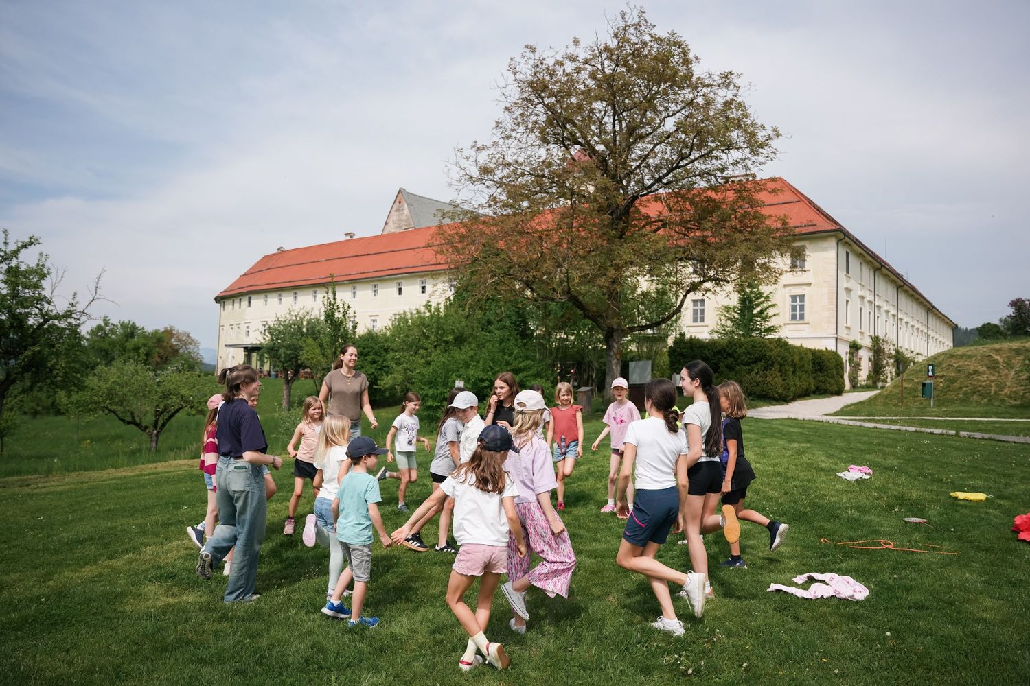 Children and adults playing a group game on green lawn outside a historic white building.