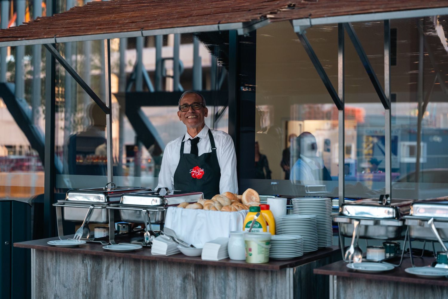 Friendly catering staff member standing behind a buffet service station with chafing dishes and fresh food.