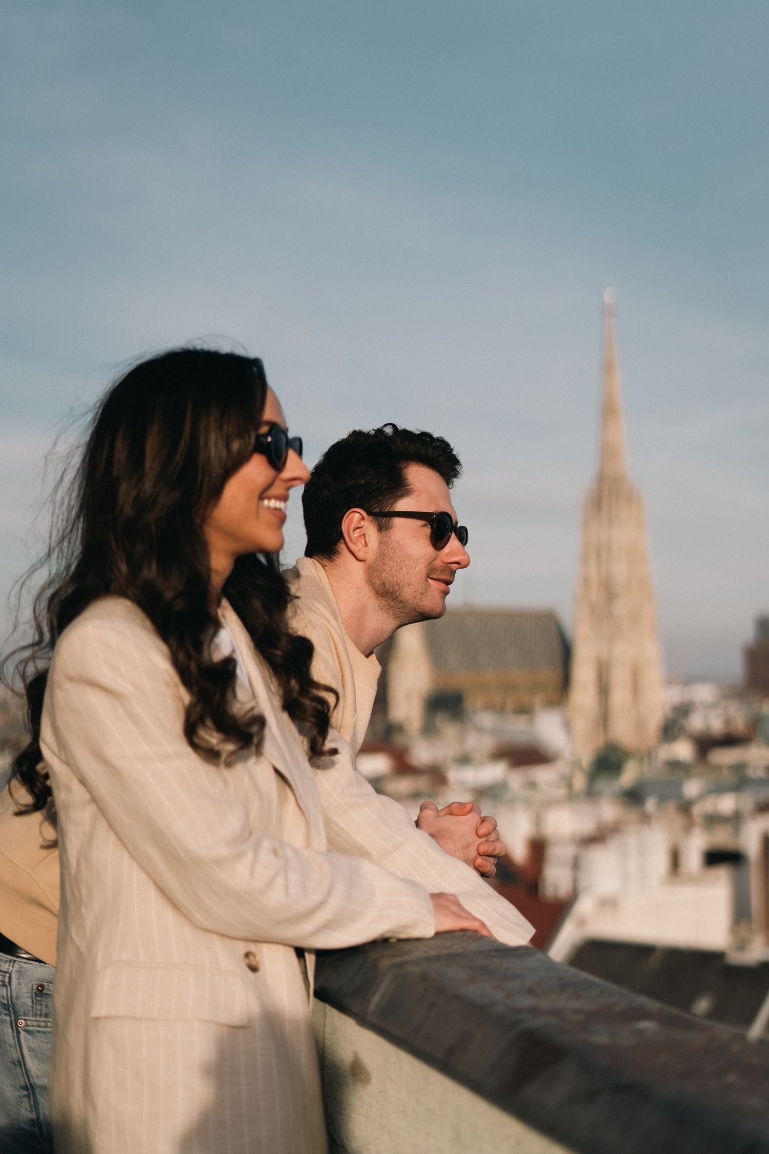 Smiling couple in sunglasses leaning on a rooftop railing with a cathedral spire behind them.