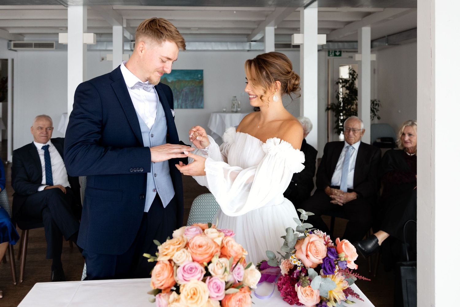 Groom placing wedding ring on brides finger at elegant ceremony with colorful floral arrangement.