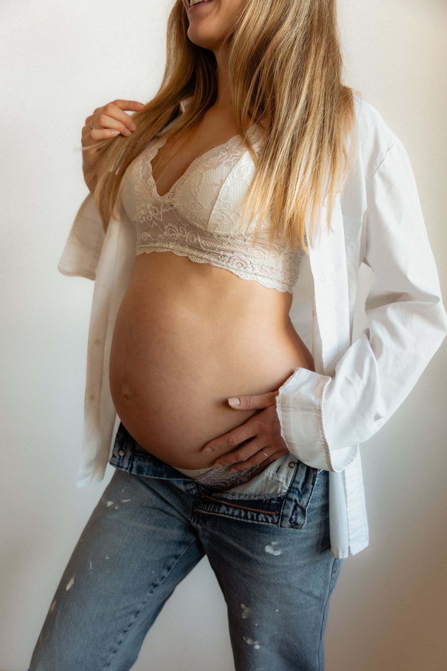 Pregnant woman in open white shirt and lace bralette gently holding her exposed baby bump.