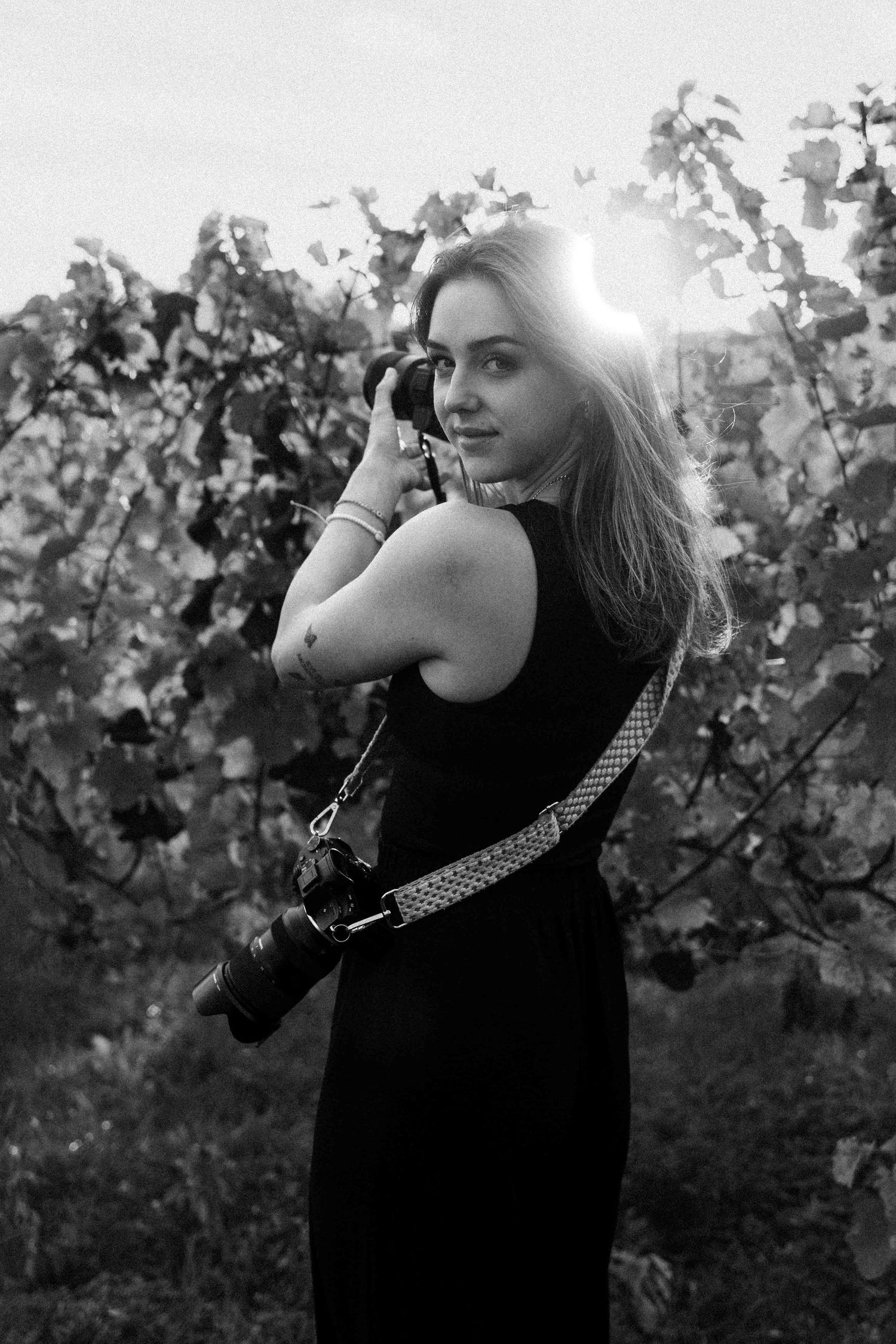 Black and white portrait of a female photographer posing in a sunlit vineyard with camera.