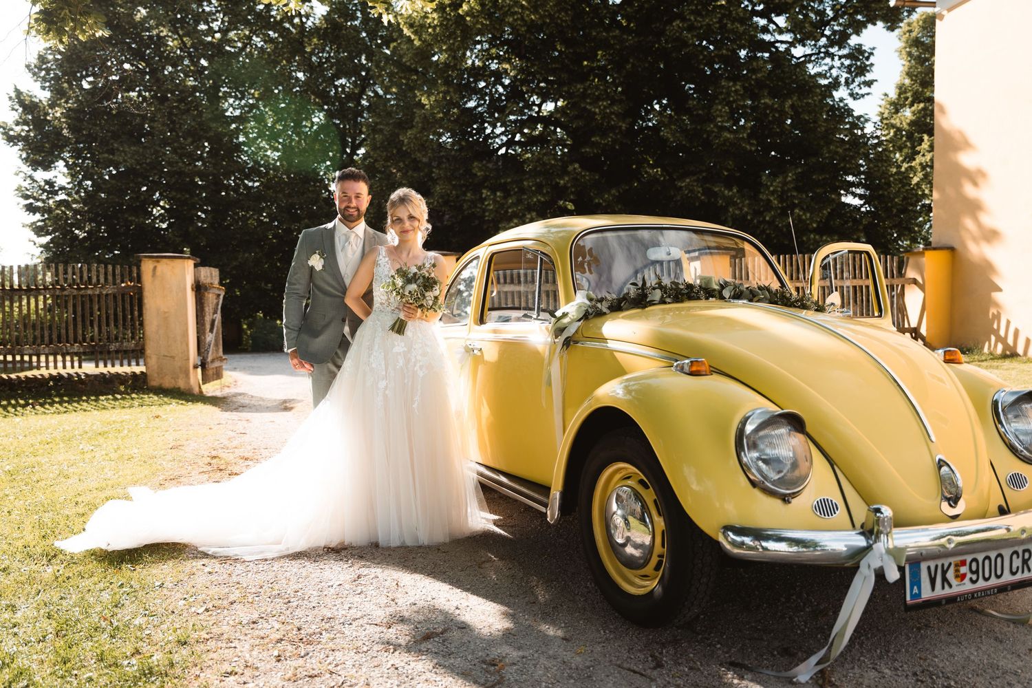 Bride and groom posing beside a classic yellow Volkswagen Beetle decorated with wedding ribbons outdoors.