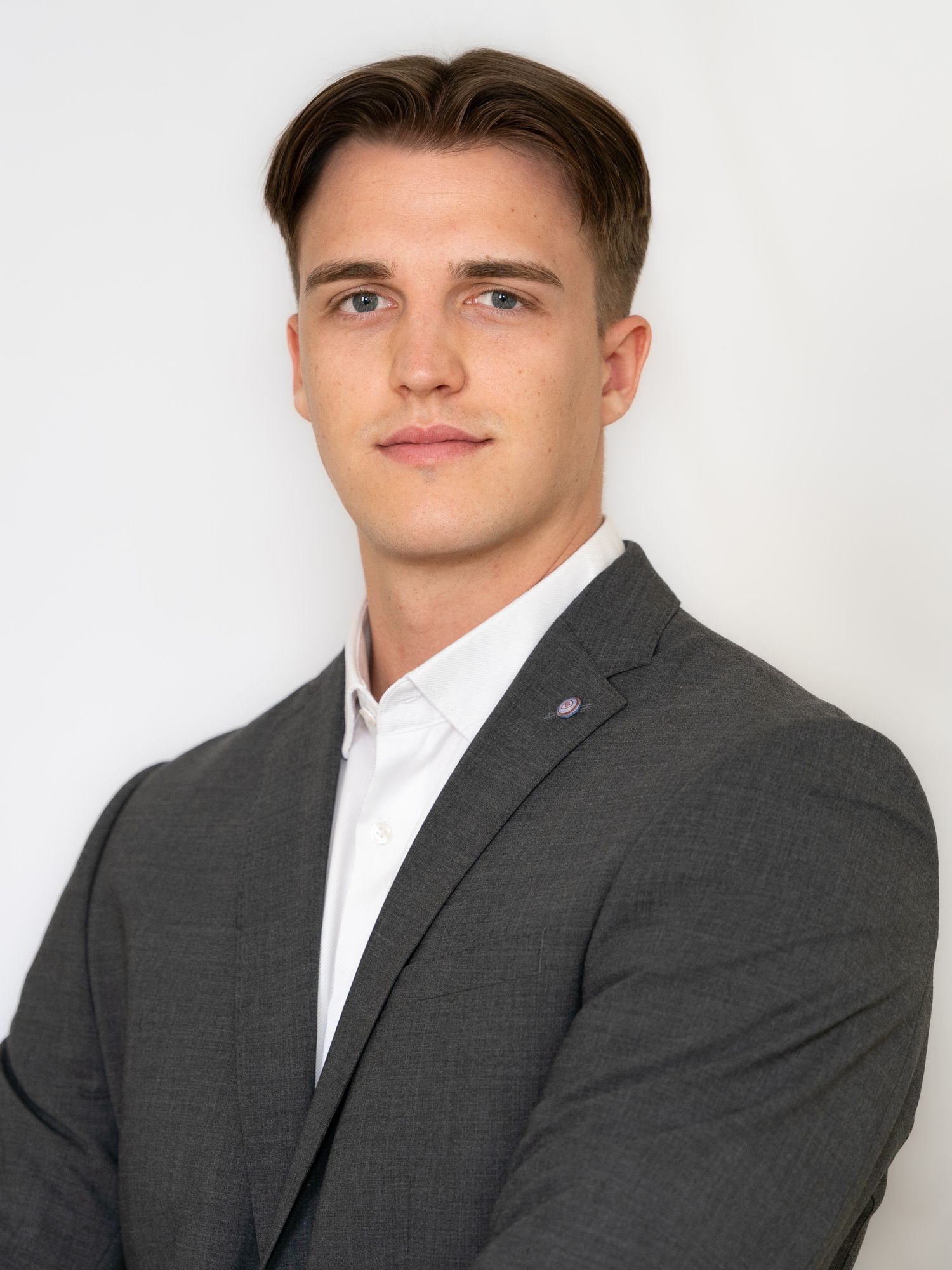 Professional headshot of a young man in a gray blazer and white shirt against a white background.