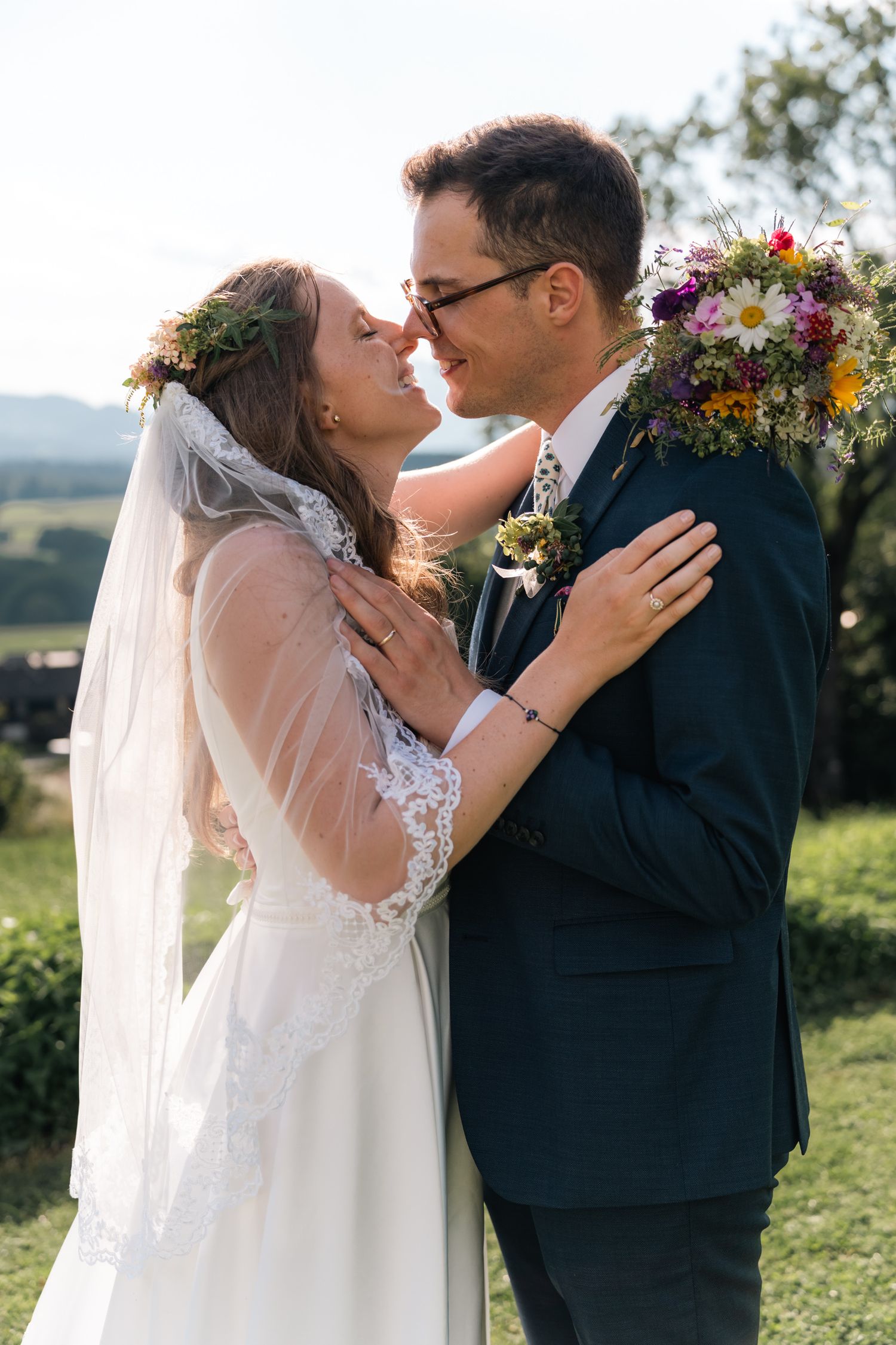 Bride and groom share a romantic almost-kiss outdoors, bride wearing floral crown and veil, groom in navy suit with wildflower boutonniere.