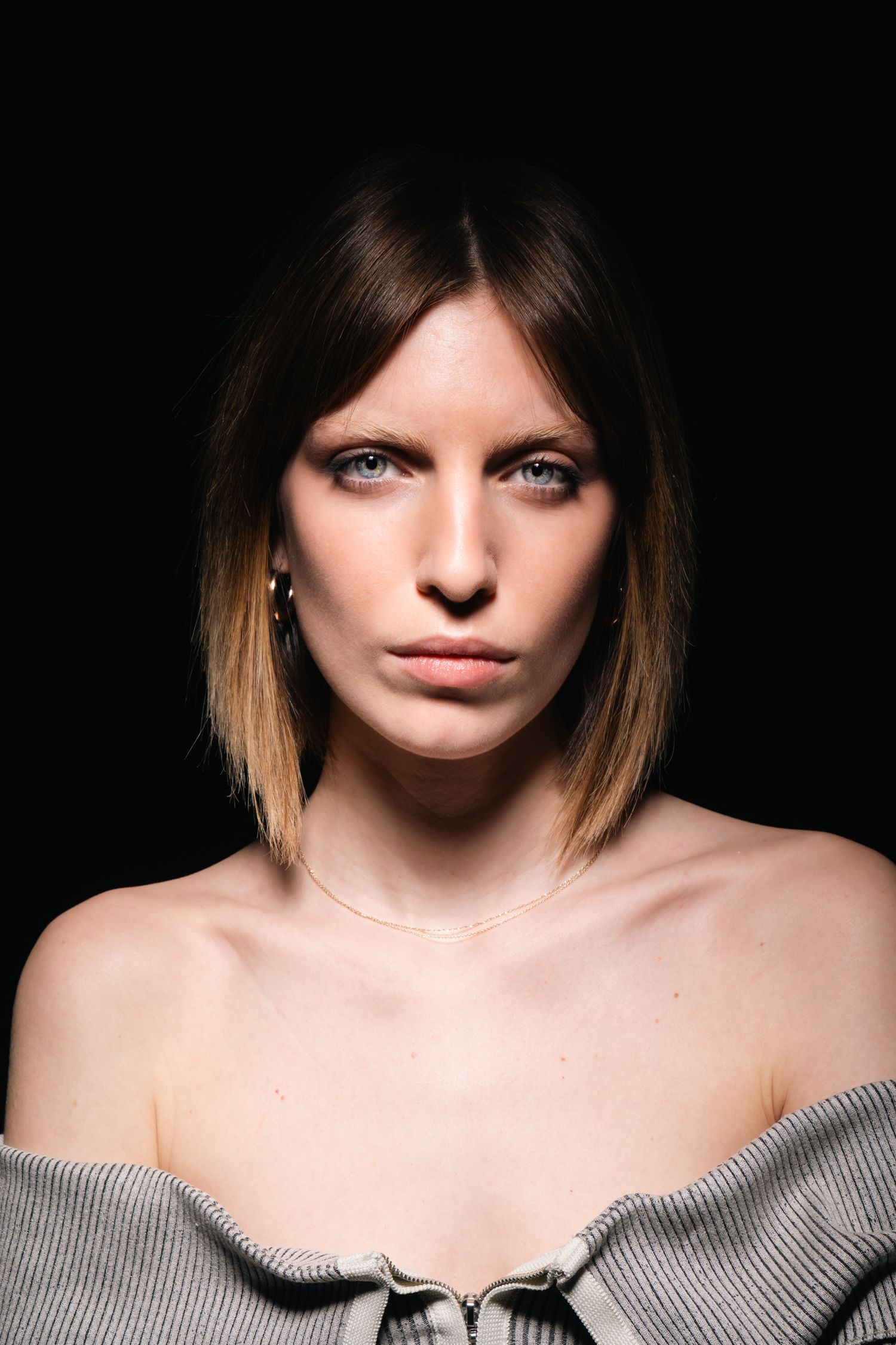 Close-up portrait of a woman with a dark blonde bob haircut wearing an off-shoulder gray top on black background.