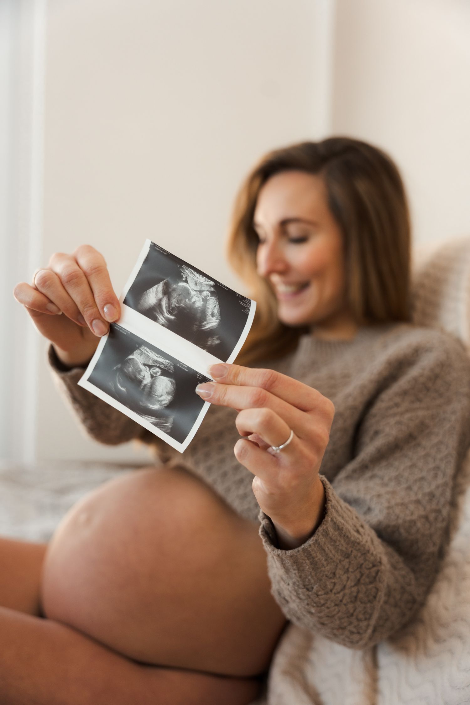 Smiling pregnant woman in a grey sweater happily looking at ultrasound photos of her baby.