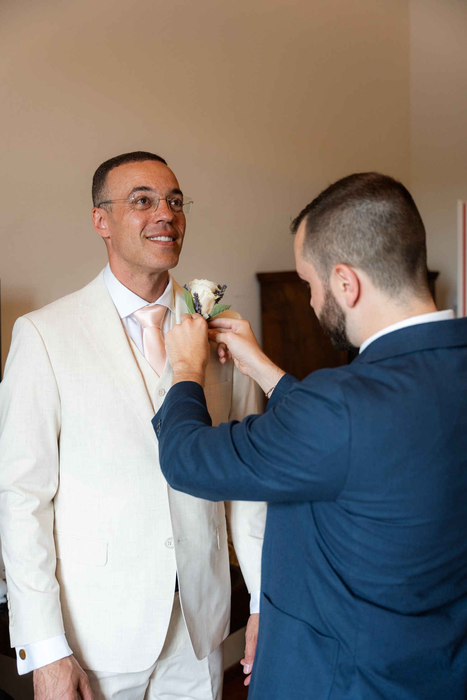 Groomsman in navy suit pinning a white boutonniere on smiling groom in cream suit.