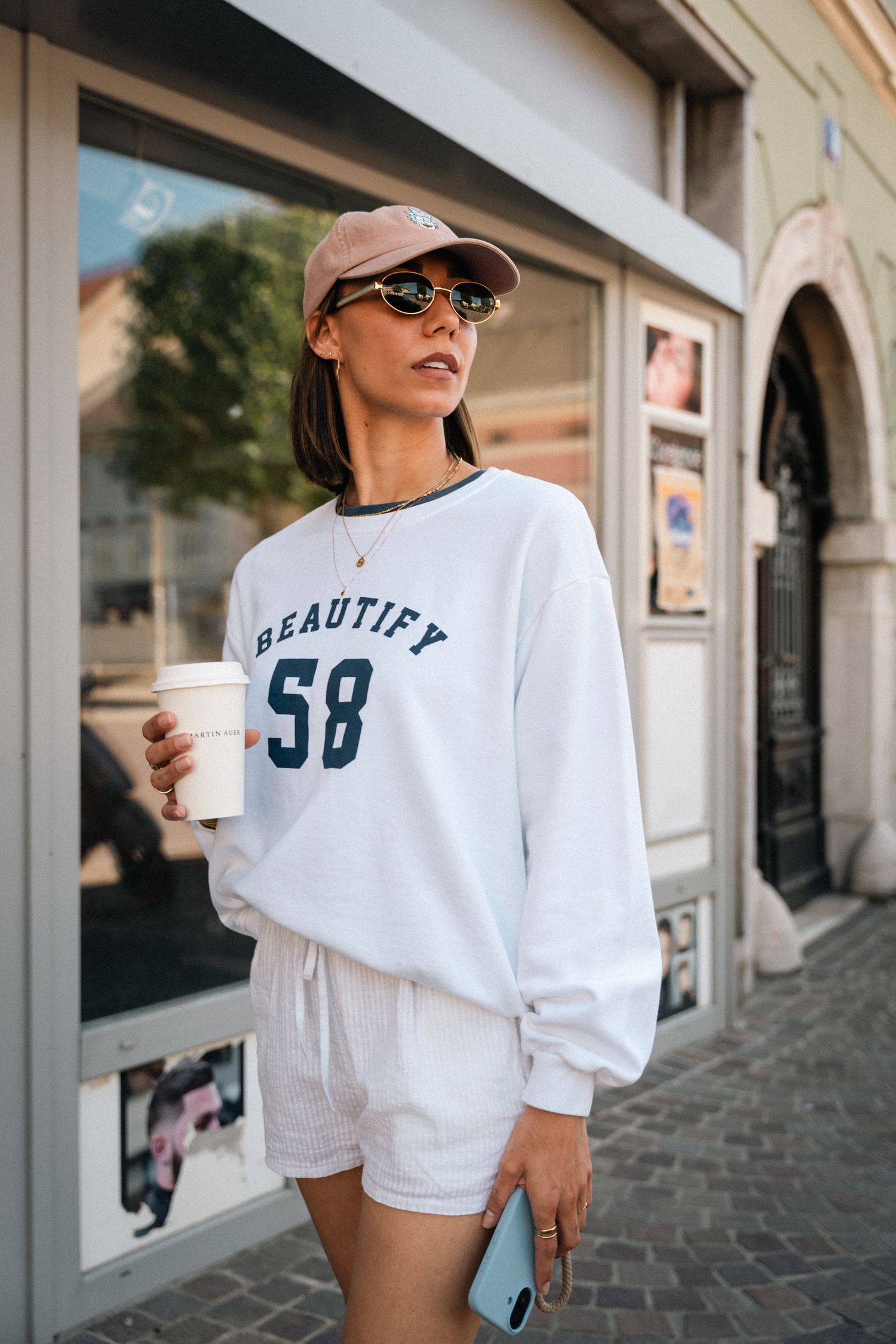 Woman in white Beautify 58 sweatshirt holding coffee cup on city street.