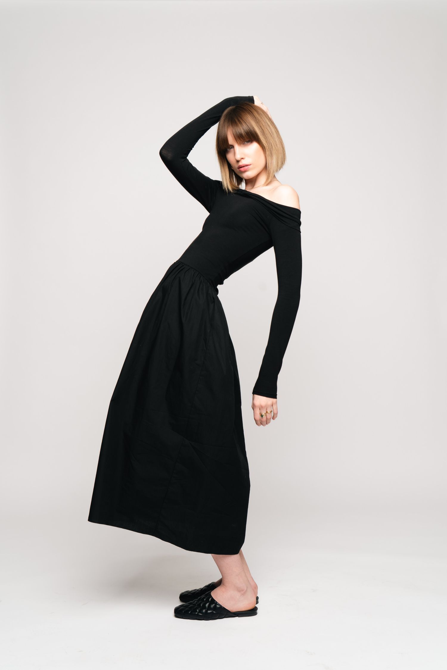 Woman posing in an elegant off-shoulder black maxi dress with studded mules in a studio.