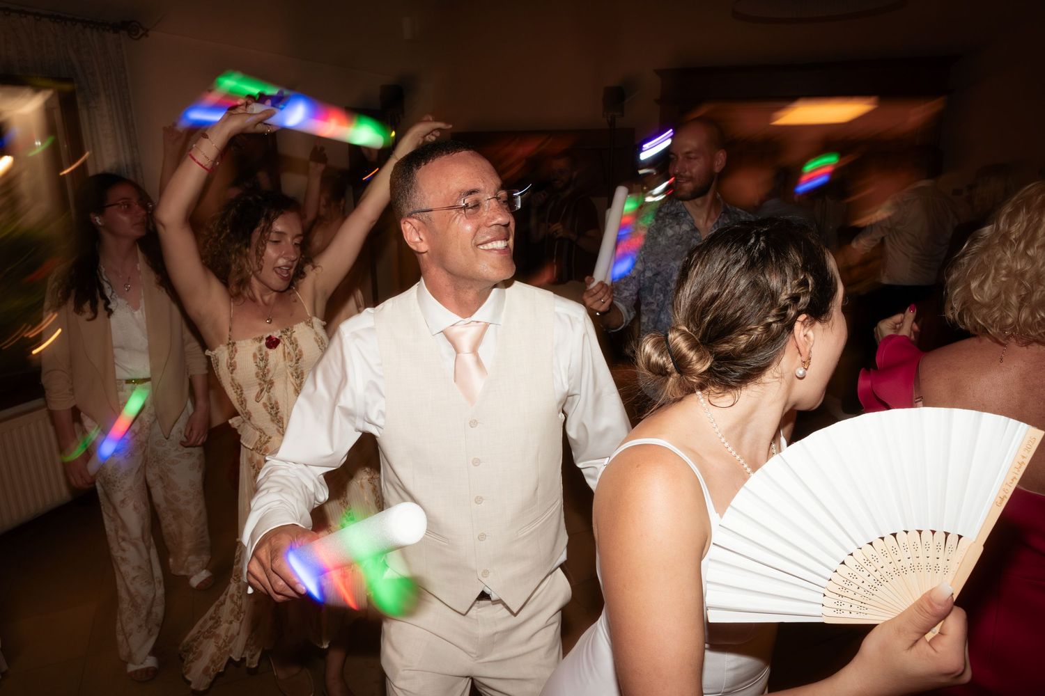 Wedding guests dance joyfully with colorful glow sticks at a lively indoor wedding reception party.