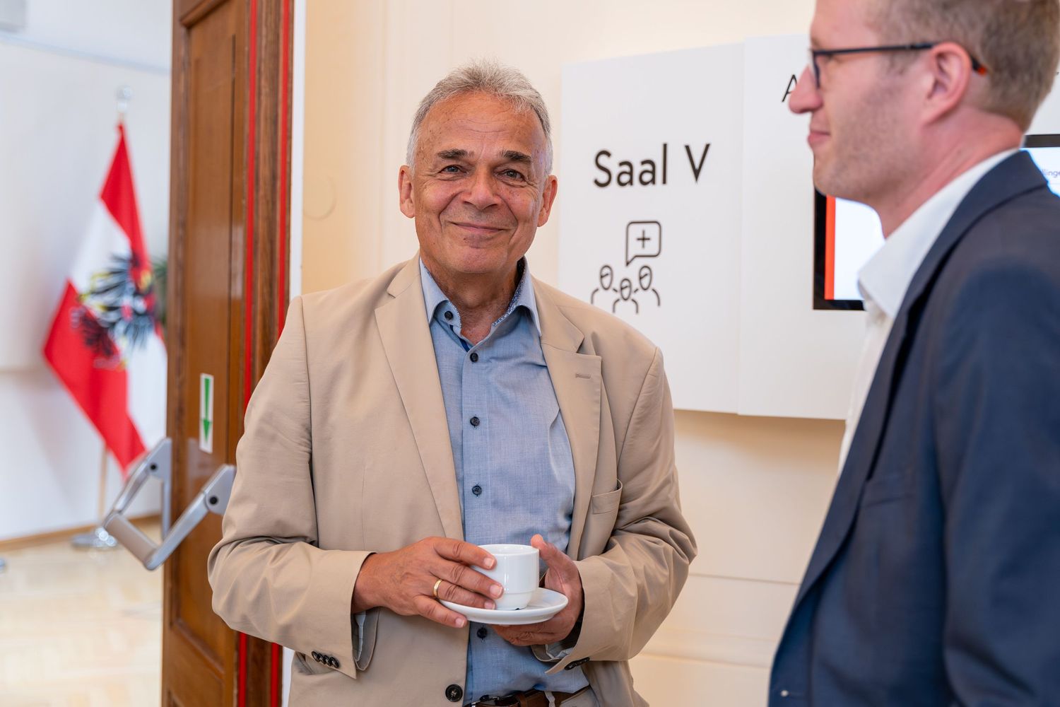 Two men networking at a professional event near a door labeled Saal V with an Austrian flag in background.
