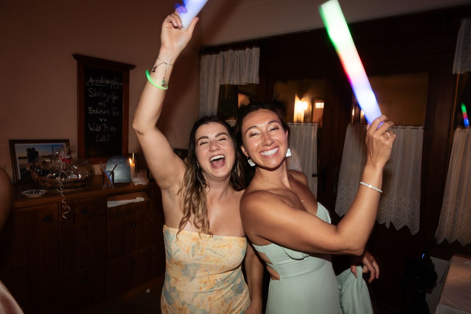Two bridesmaids in pastel gowns dancing joyfully with colorful glow sticks at wedding reception.