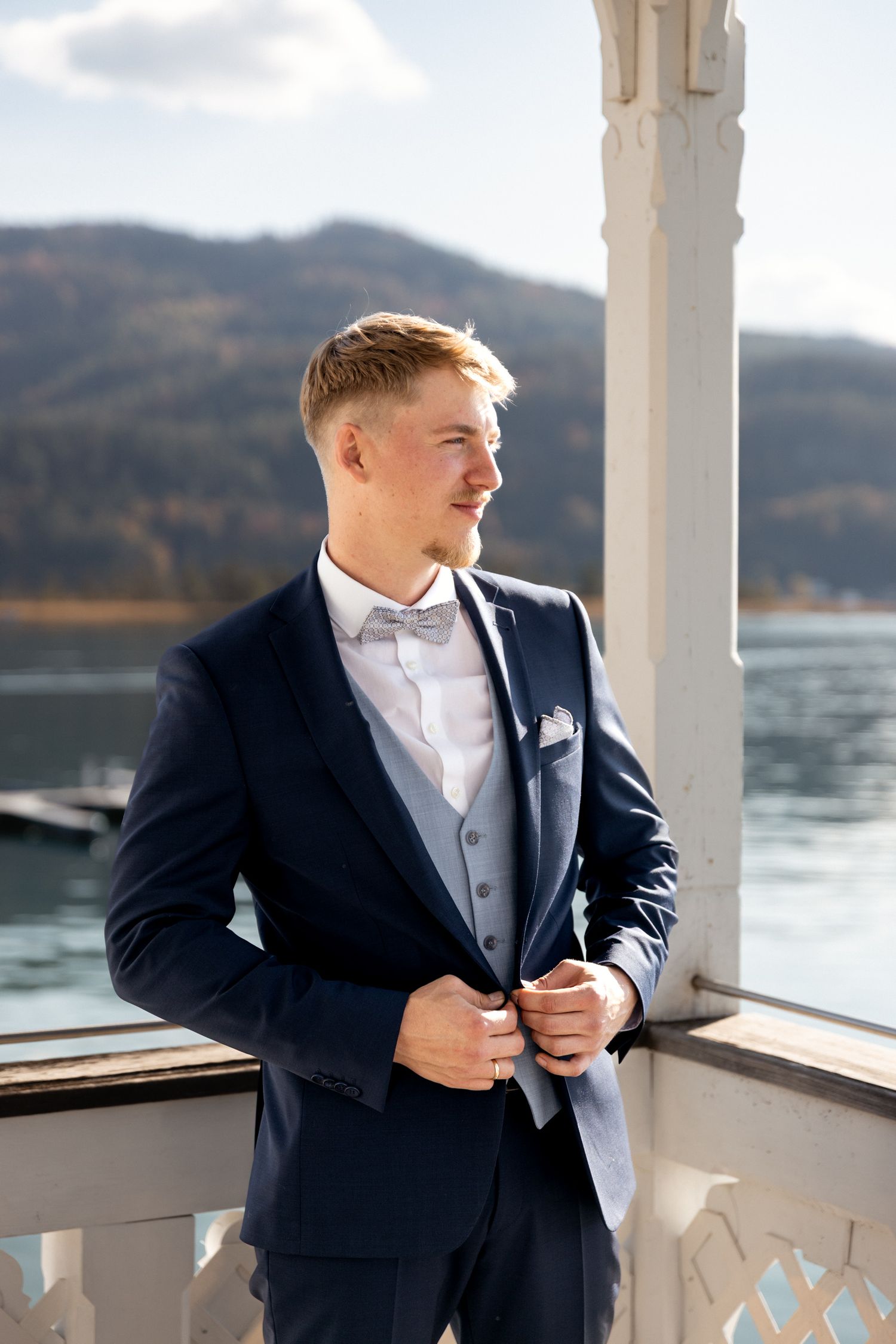 Groom in navy suit with bow tie poses by waterfront on wedding day.