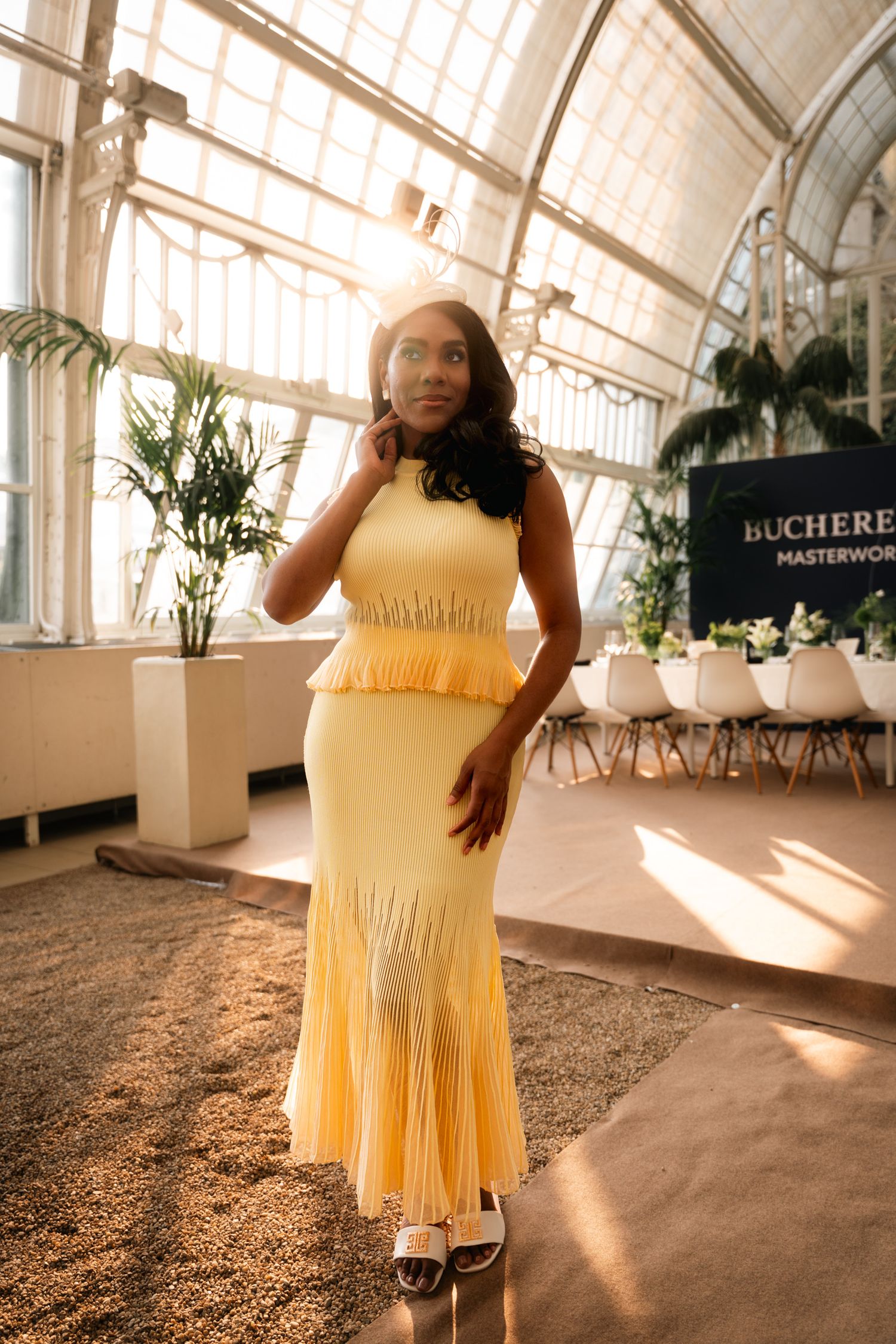 Woman in yellow pleated midi dress posing in sunlit greenhouse venue.