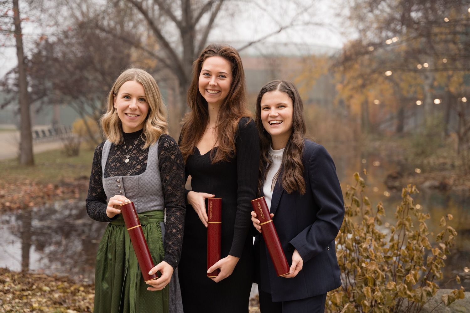 Three young women in formal attire holding diploma tubes at outdoor graduation ceremony.