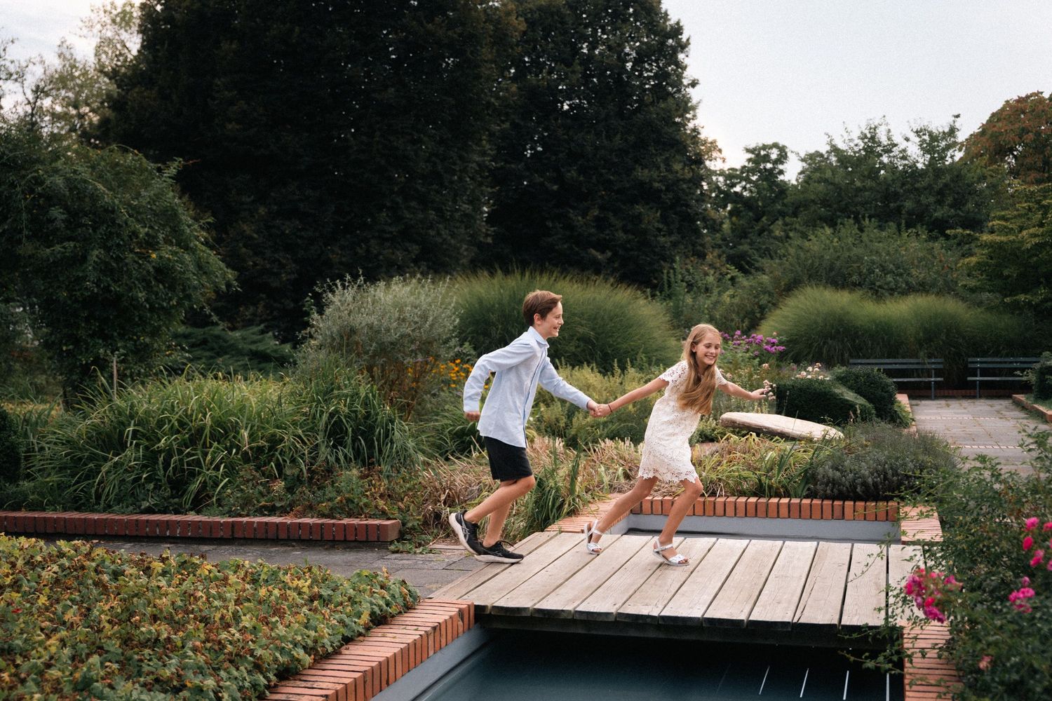 A joyful couple runs hand-in-hand across a wooden bridge in a lush green garden.