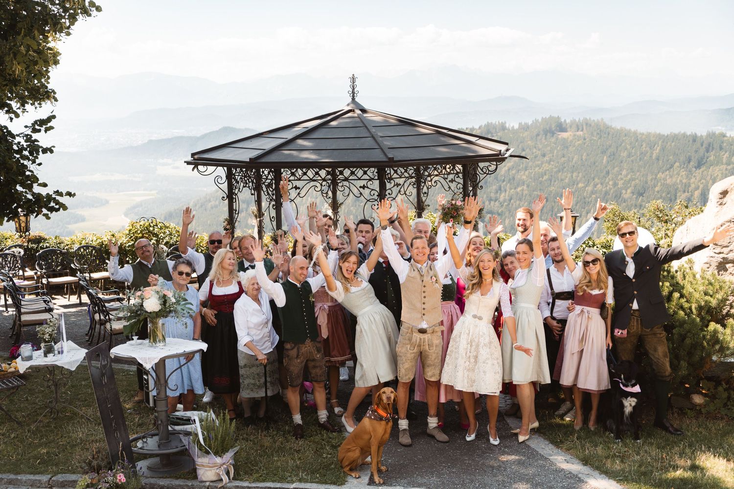 Wedding guests in traditional Austrian dirndl and lederhosen celebrating with raised hands at scenic overlook.