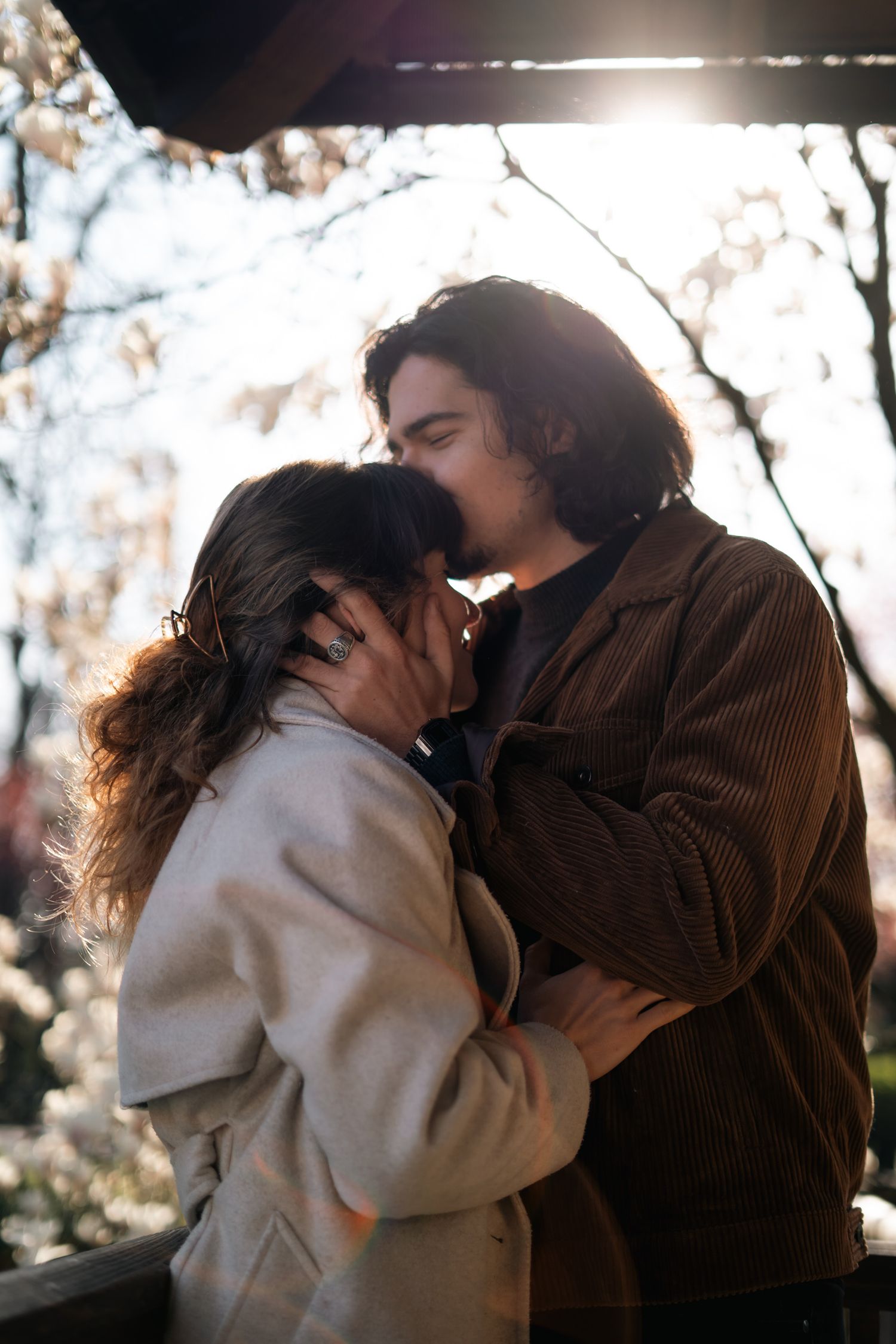 Couple sharing a tender kiss outdoors with sunlight filtering through blossoming trees behind them.