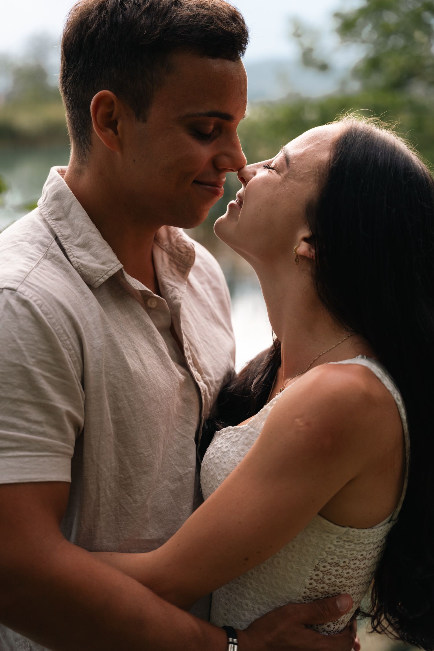 Young couple in intimate moment, nearly kissing outdoors near water with soft natural lighting.