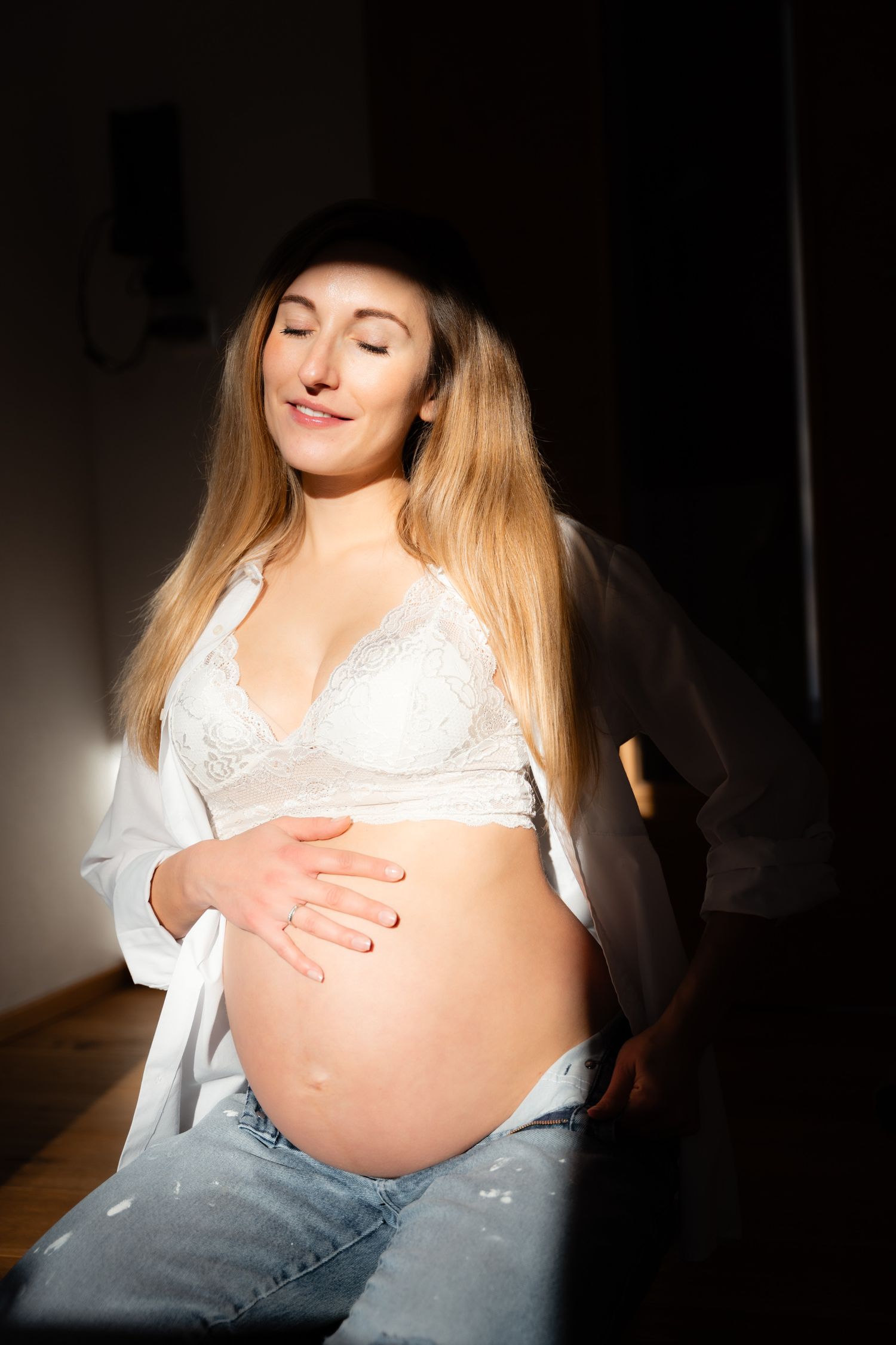 Serene pregnant woman in white lace bra and jeans with eyes closed enjoying warm sunlight.