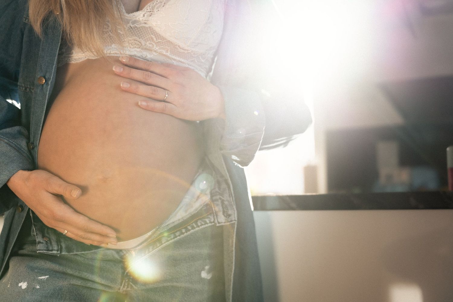 Close-up of pregnant woman in white lace bra and denim jacket holding baby bump in bright sunlight.