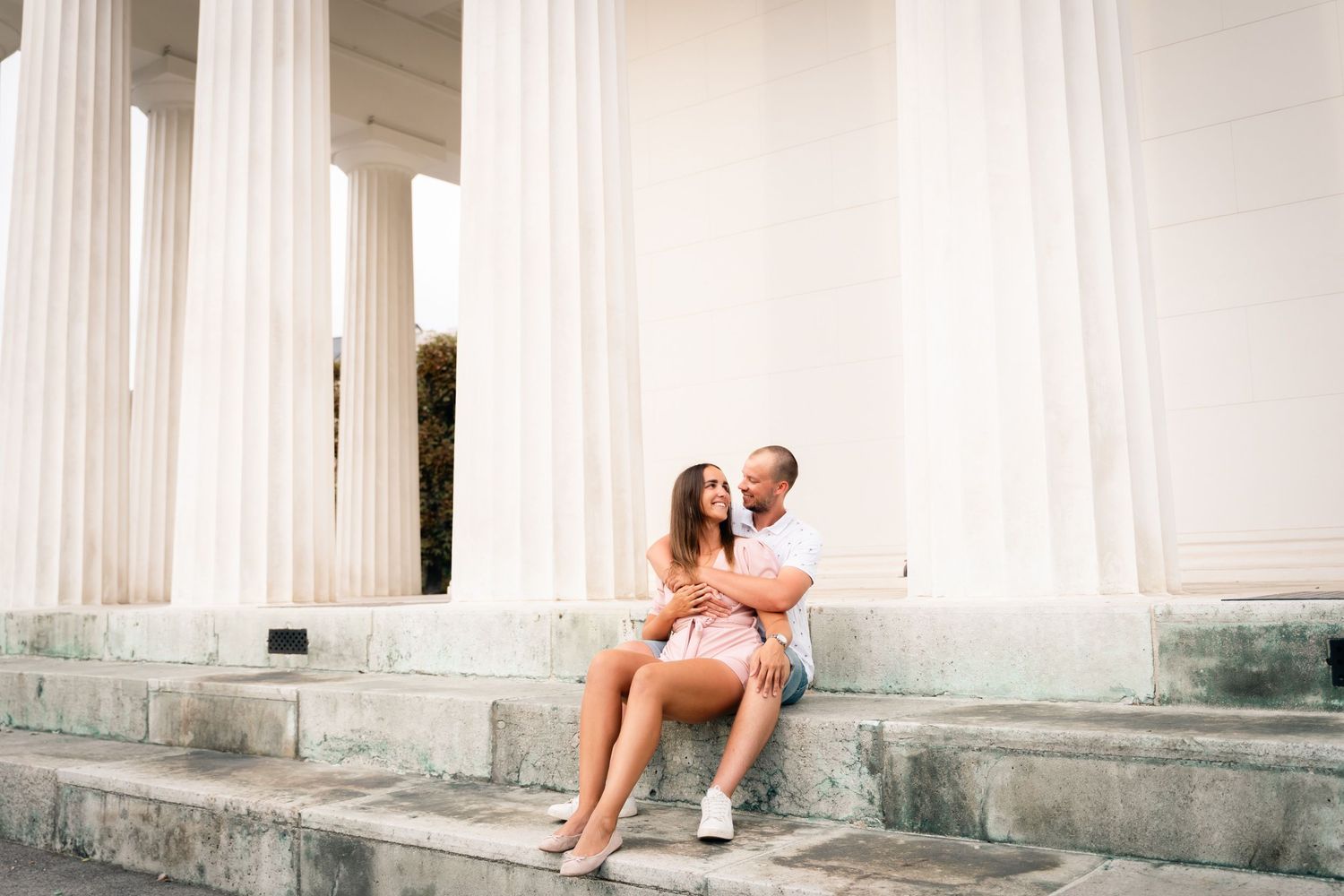 Romantic couple sitting and cuddling on steps between tall white classical columns outdoors.