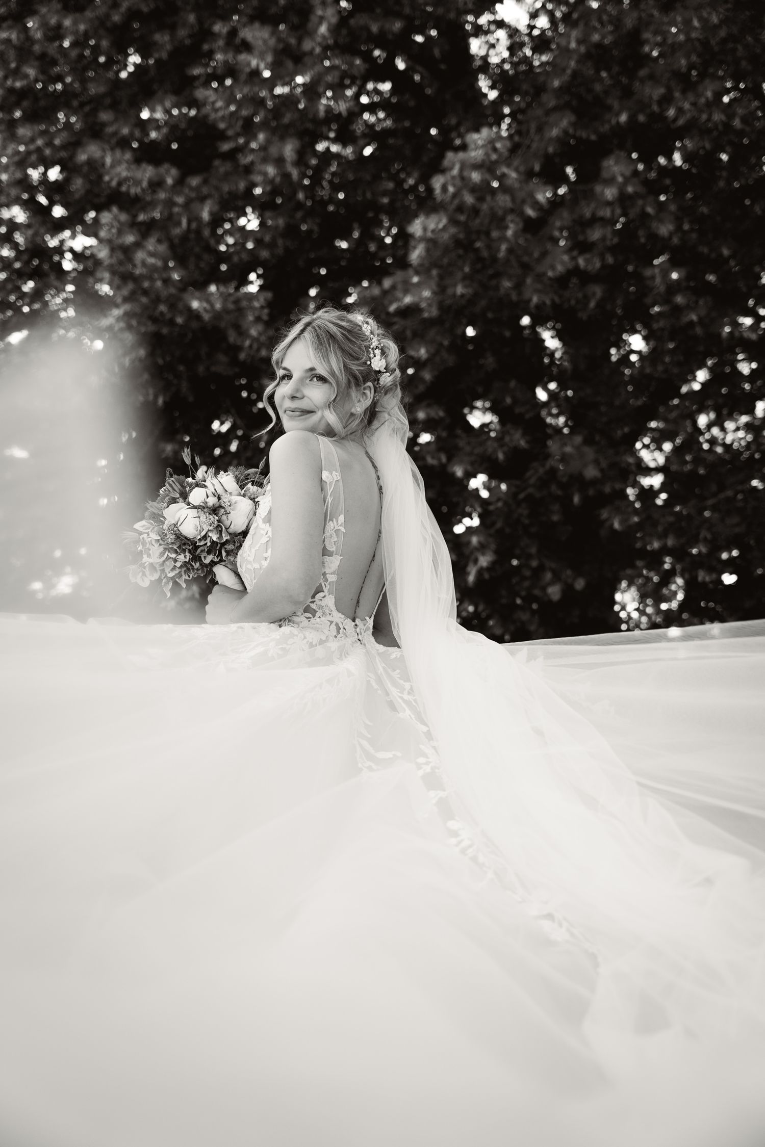 Black and white bridal portrait of a bride in a lace gown with a flowing veil holding a bouquet.