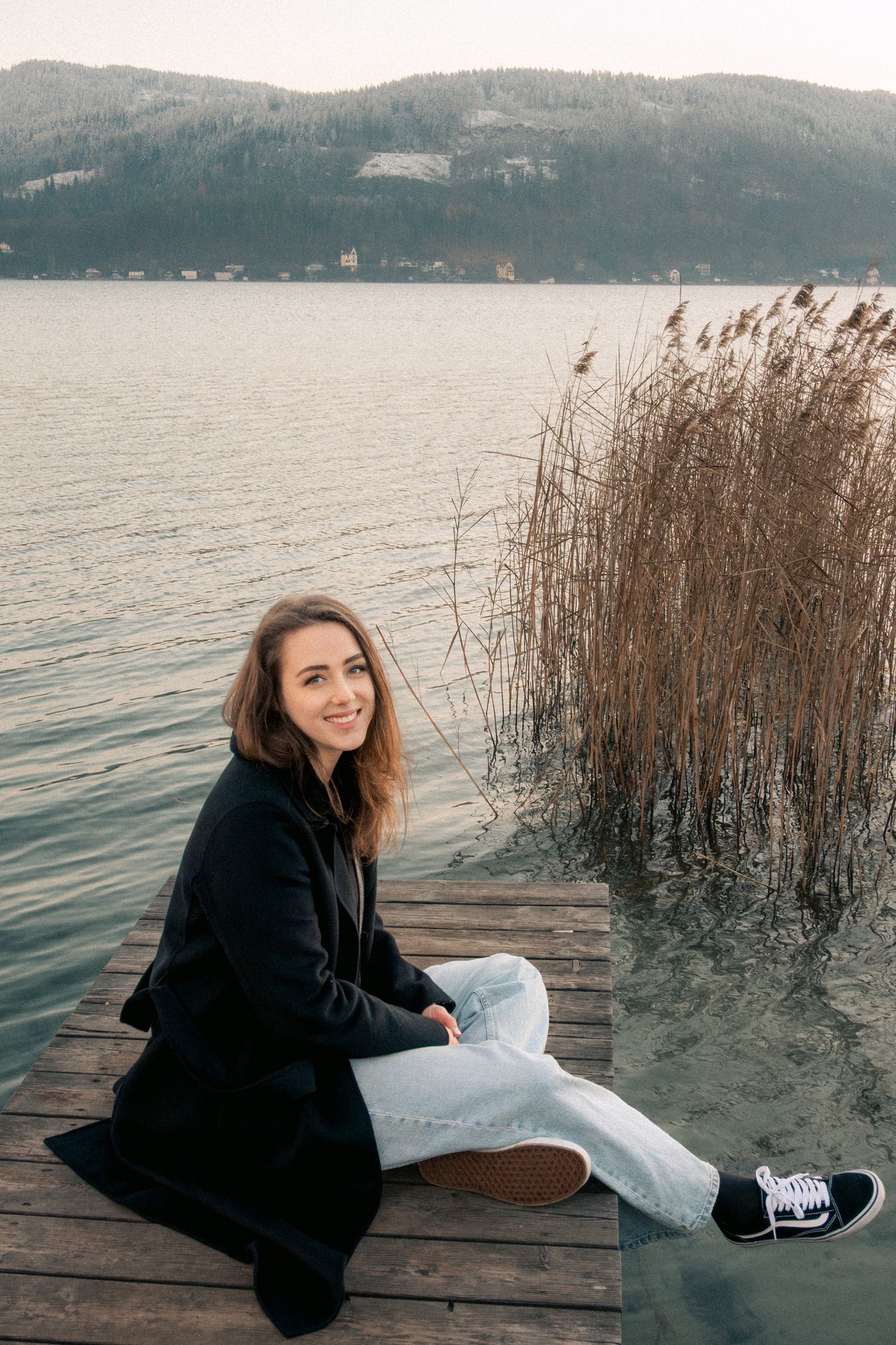 Smiling woman in black coat sitting on wooden dock by scenic mountain lake.