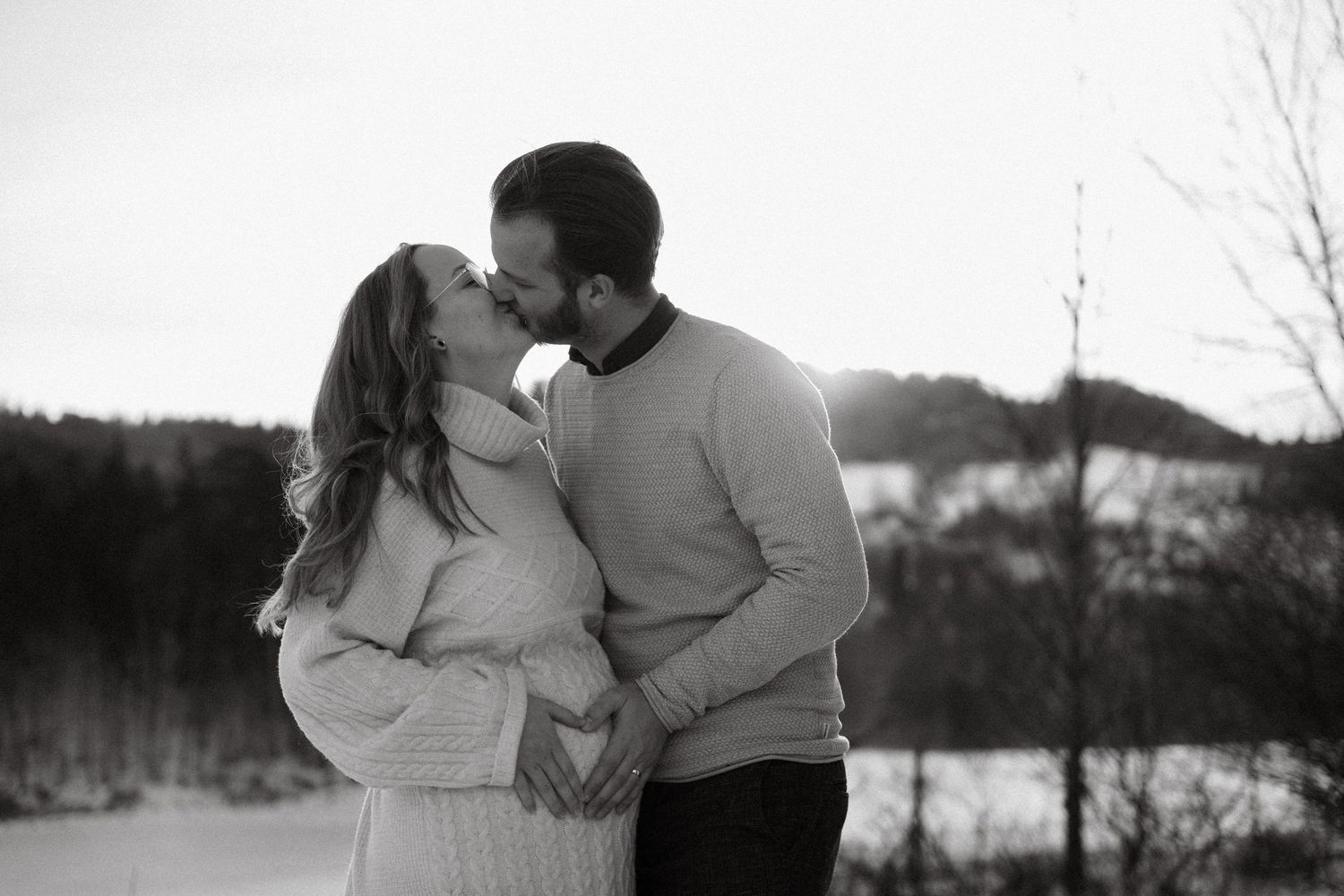 Black and white photo of couple kissing outdoors in snowy winter landscape during pregnancy.