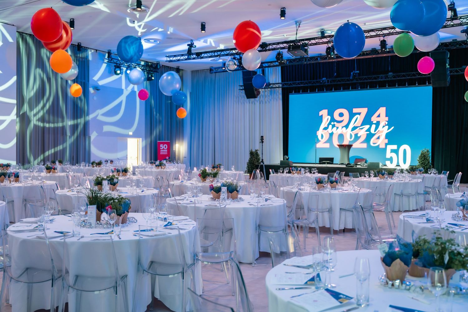 Festive 50th anniversary gala with white round tables, colorful balloons, and a large celebration screen display.