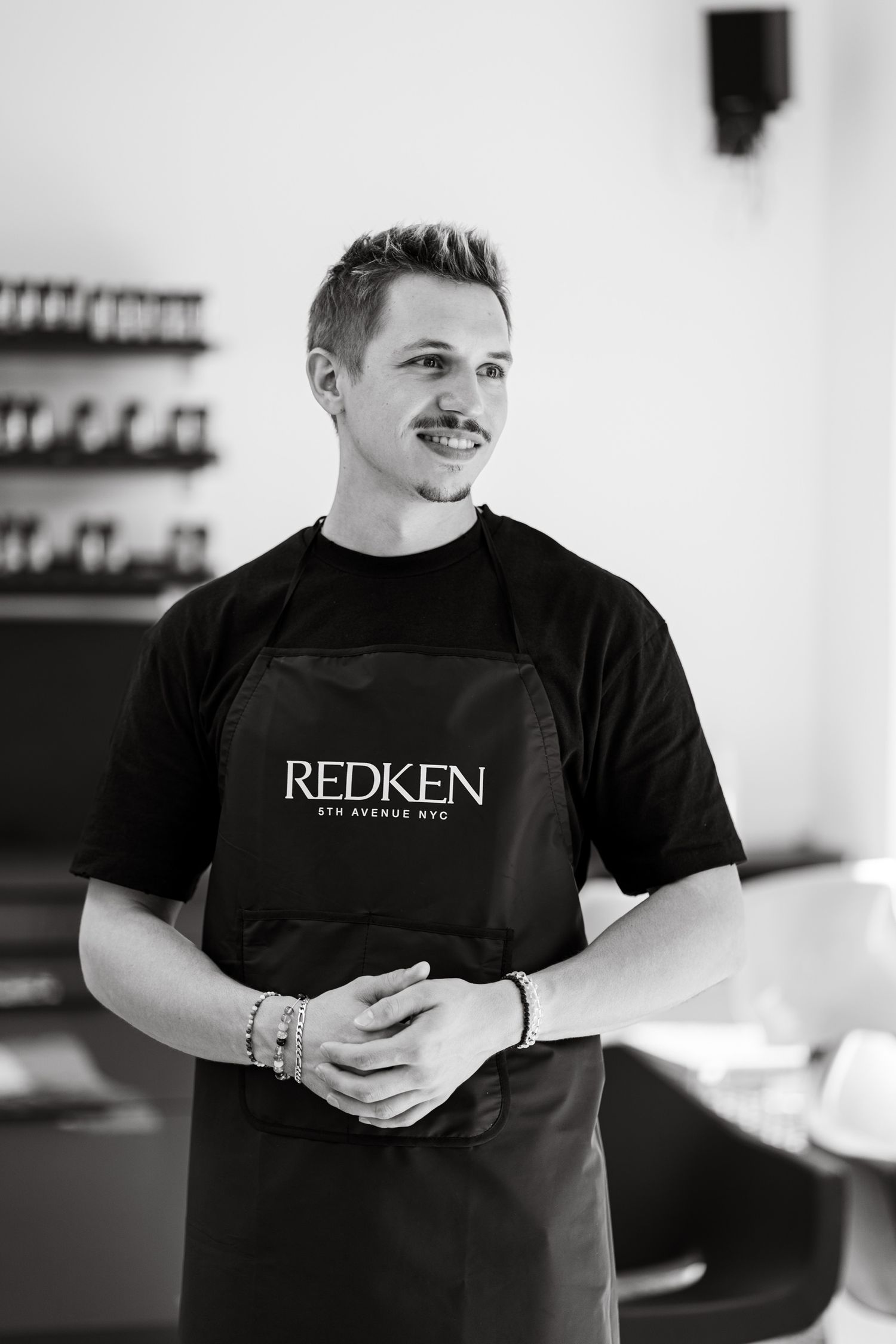 Male hairstylist in a Redken apron stands confidently in a modern hair salon.
