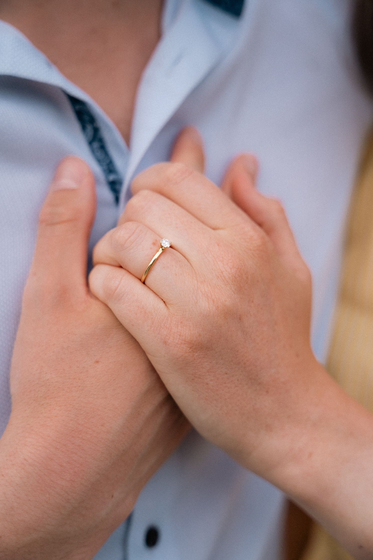 Close-up of a womans hand wearing a delicate gold solitaire engagement ring on a mans chest.