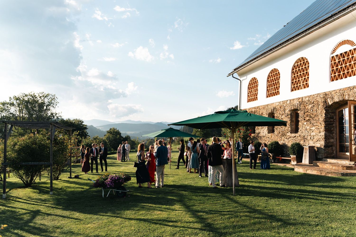 Guests mingling on a sunny green lawn beside a stone villa with mountain views in background.