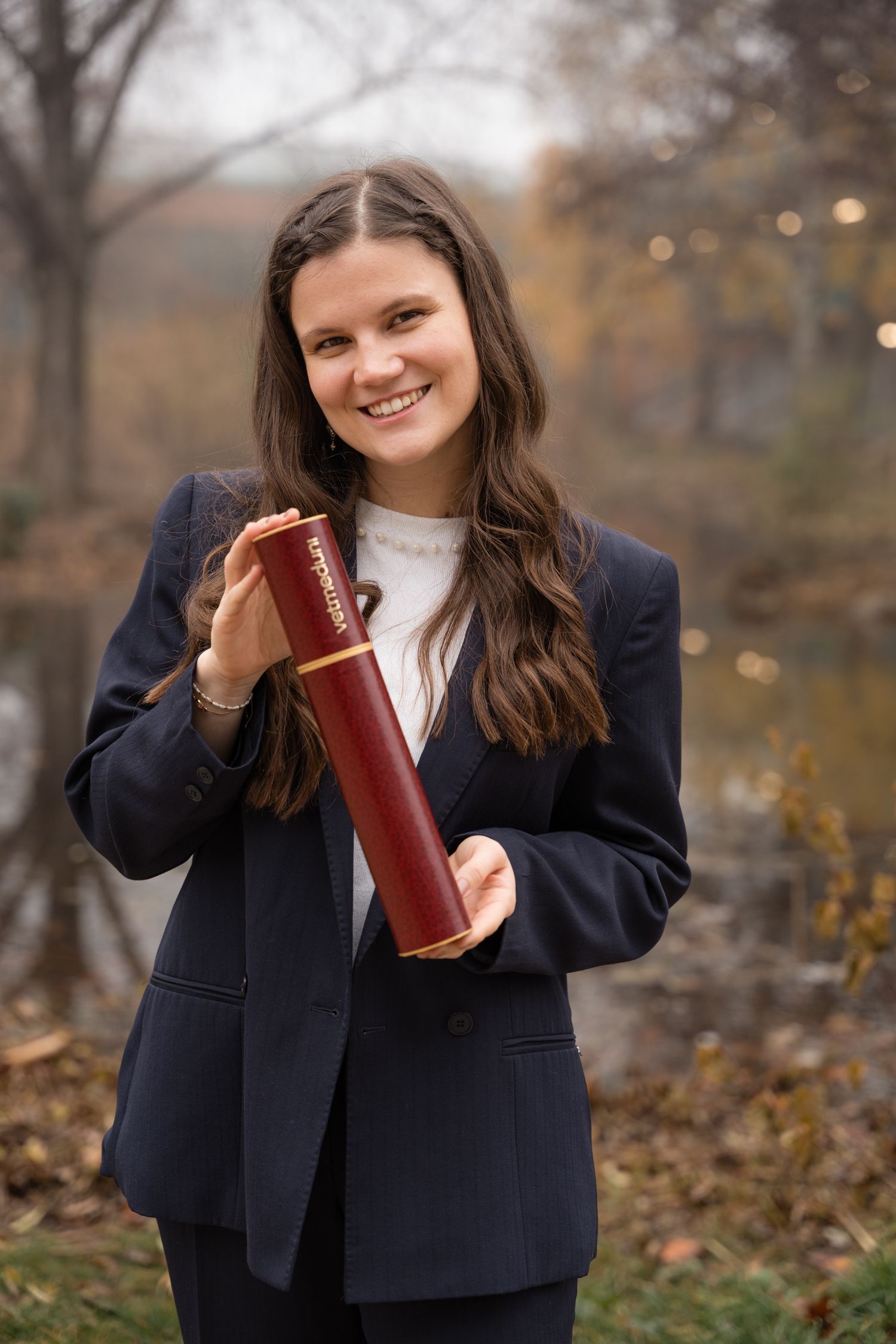 Smiling young woman in a navy blazer holding a diploma tube outdoors in an autumn setting.