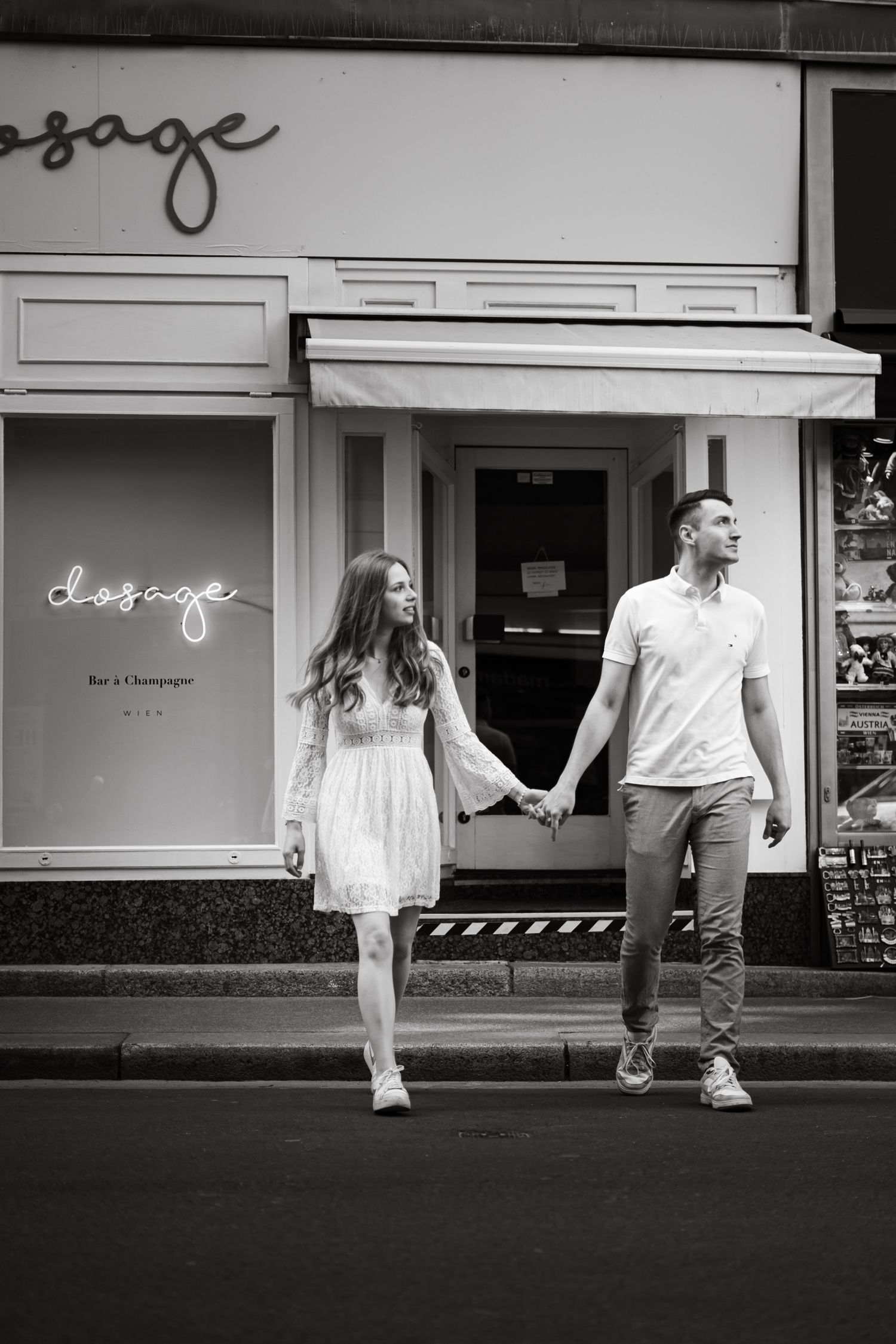 Black and white photo of a couple holding hands walking past a wine bar called Dosage on a city street.