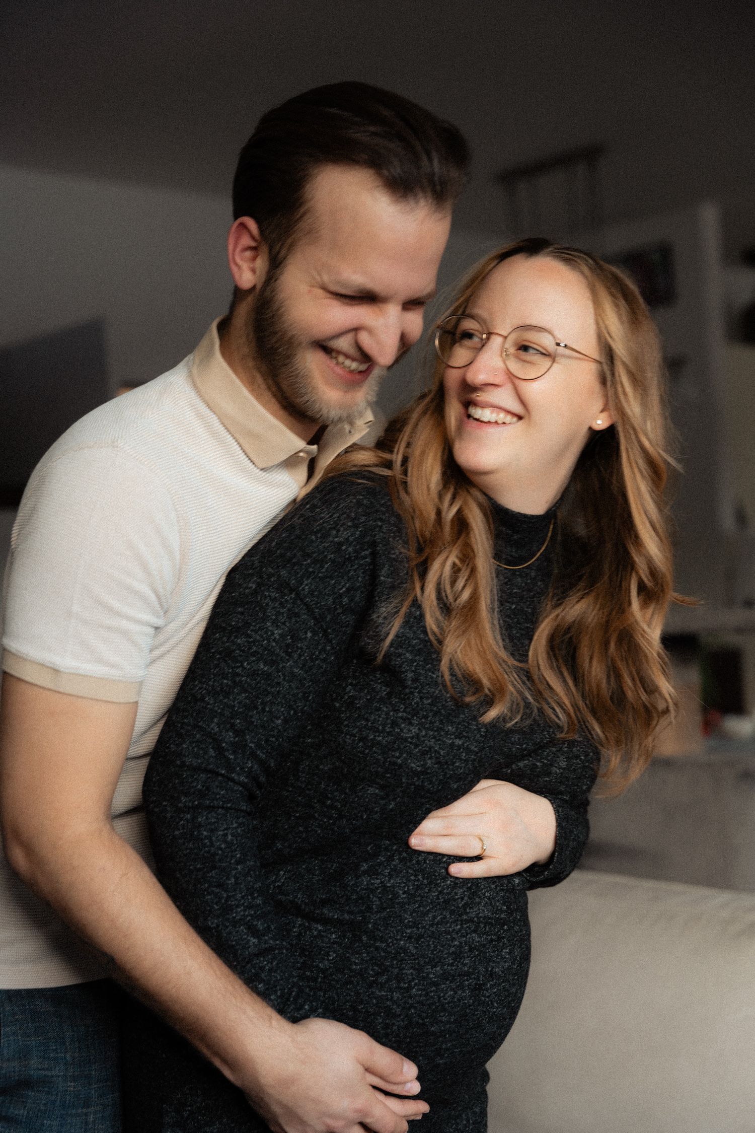Smiling couple embracing, pregnant woman in dark sweater holding baby bump indoors.