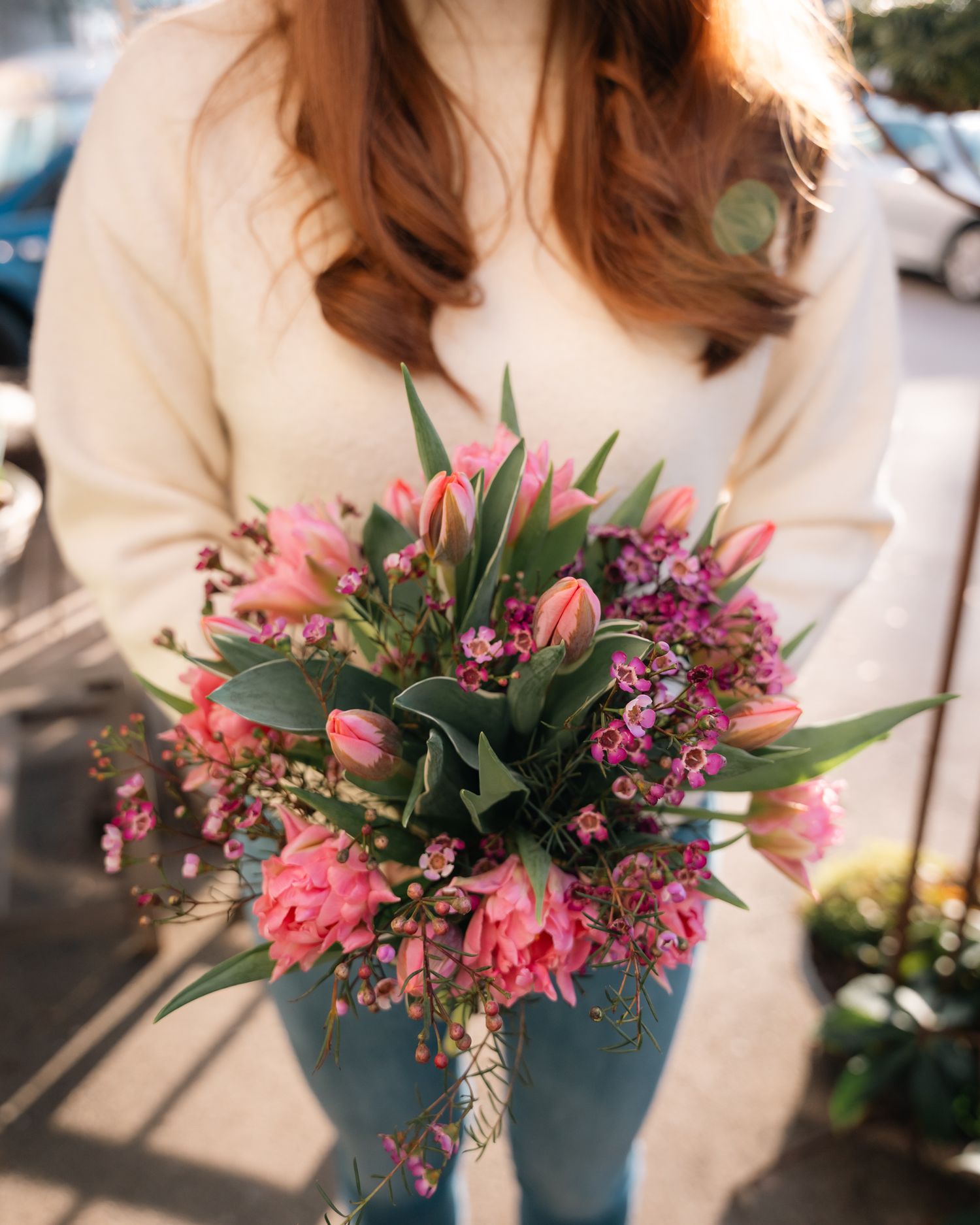 Woman in white sweater holding a vibrant bouquet of pink tulips and purple wildflowers outdoors.