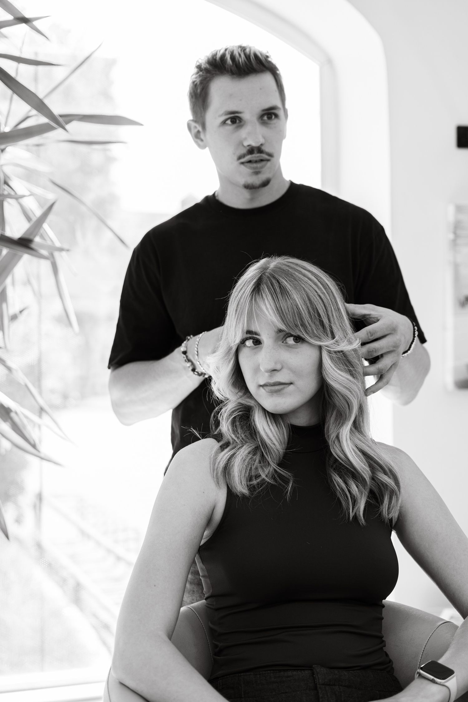 Black and white photo of a male hairstylist styling a blonde woman's hair in a modern salon.