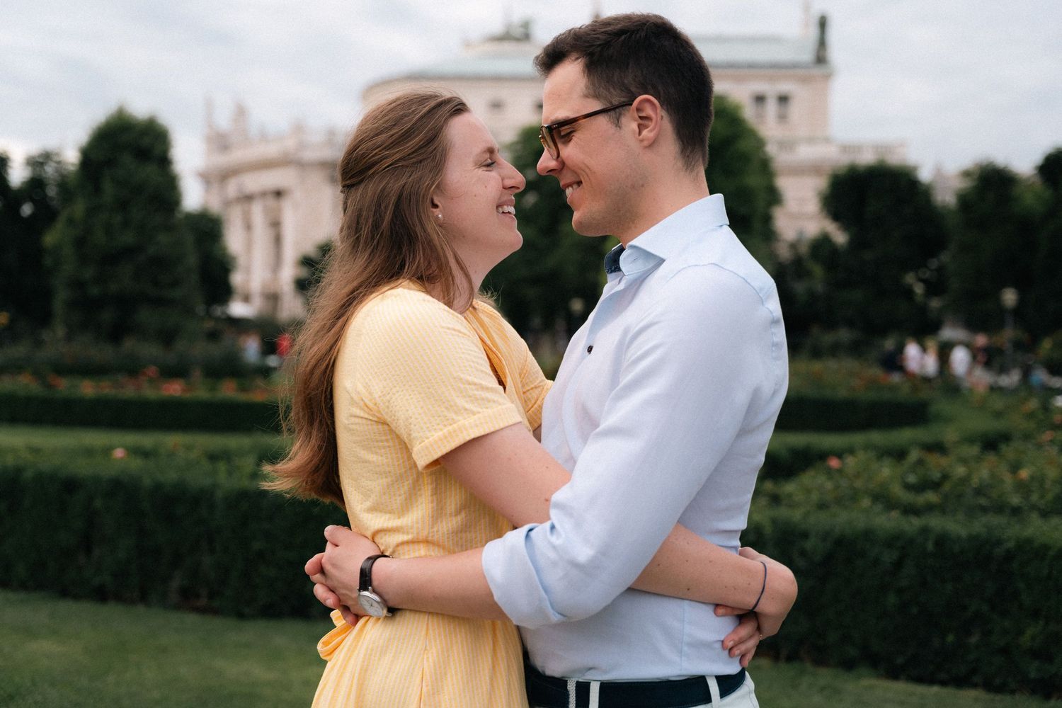 Happy couple embracing and smiling in a lush green park with a historic building in background.