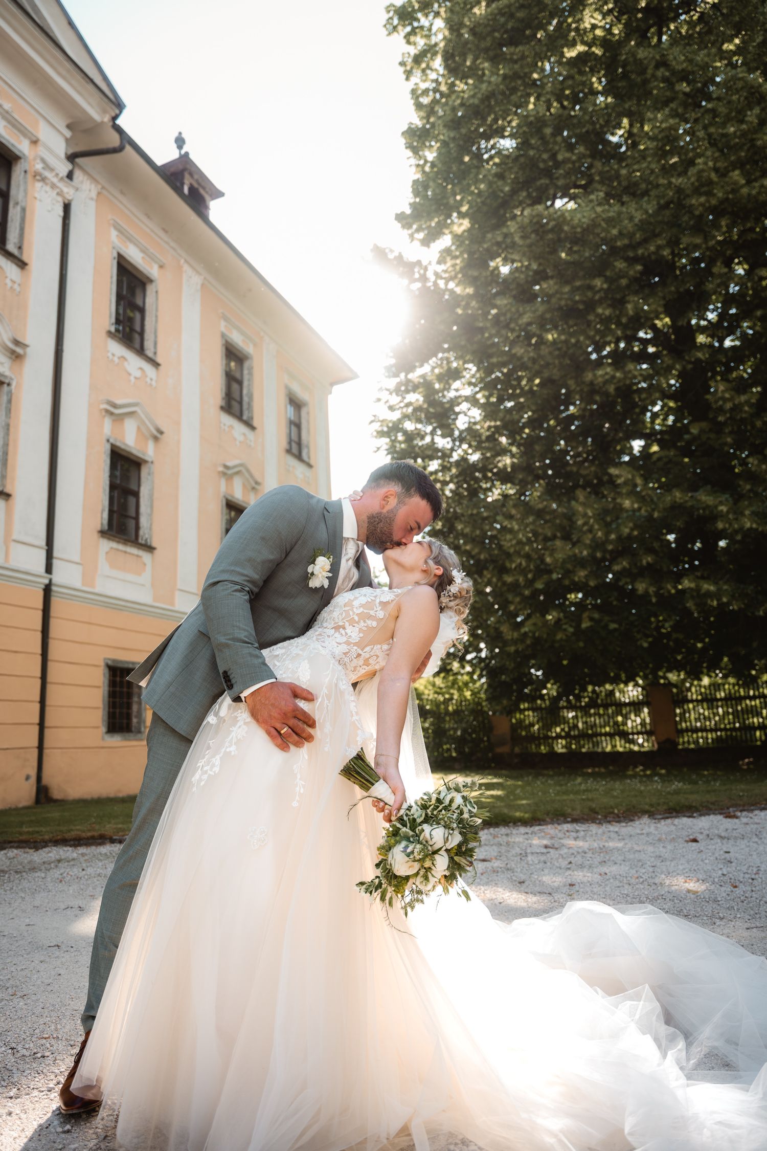 Groom in grey suit dips bride in elegant tulle gown for a romantic kiss outside a historic manor.