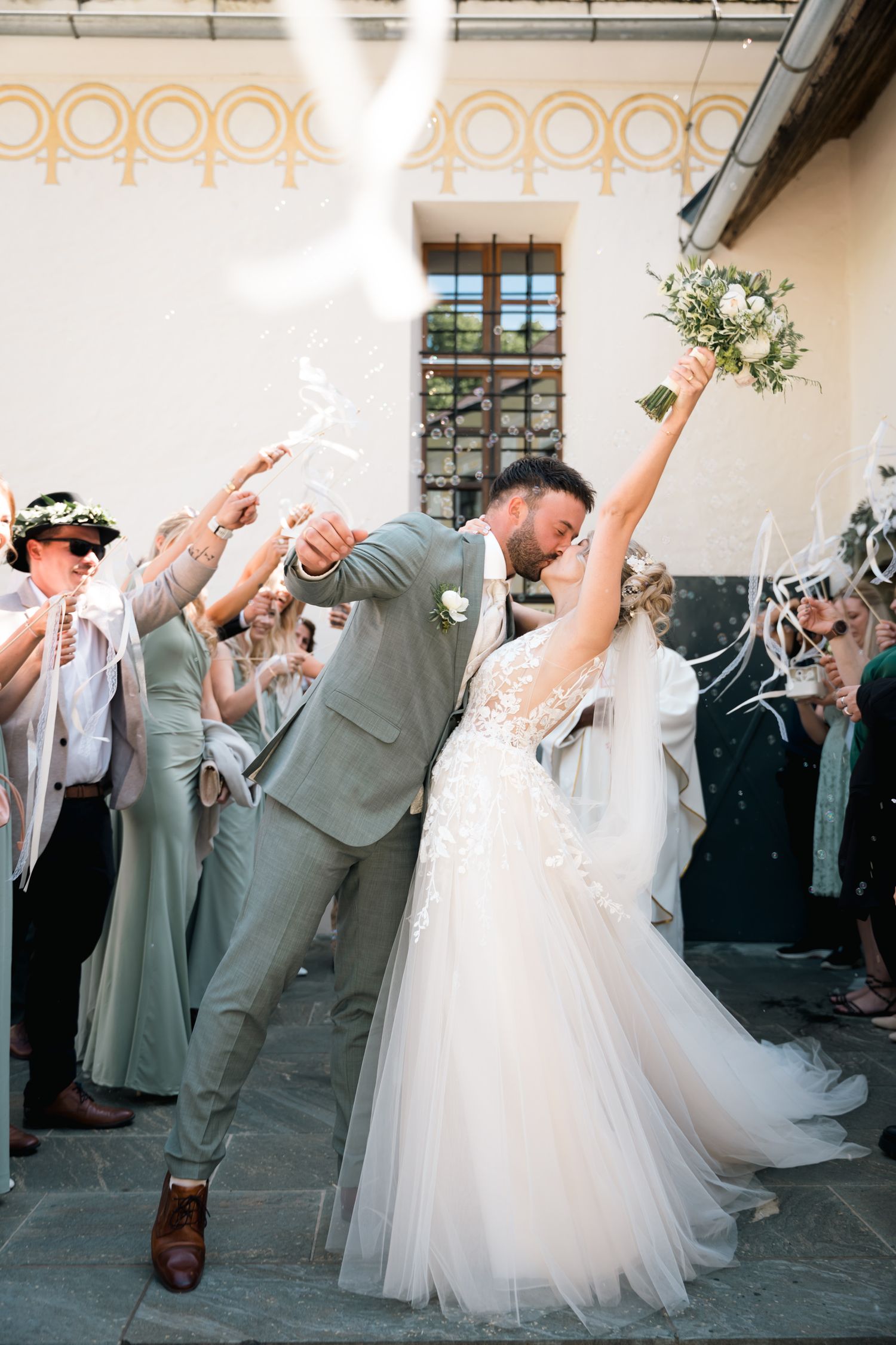 Joyful newlyweds kissing as guests celebrate with sparklers and ribbons after outdoor wedding ceremony.