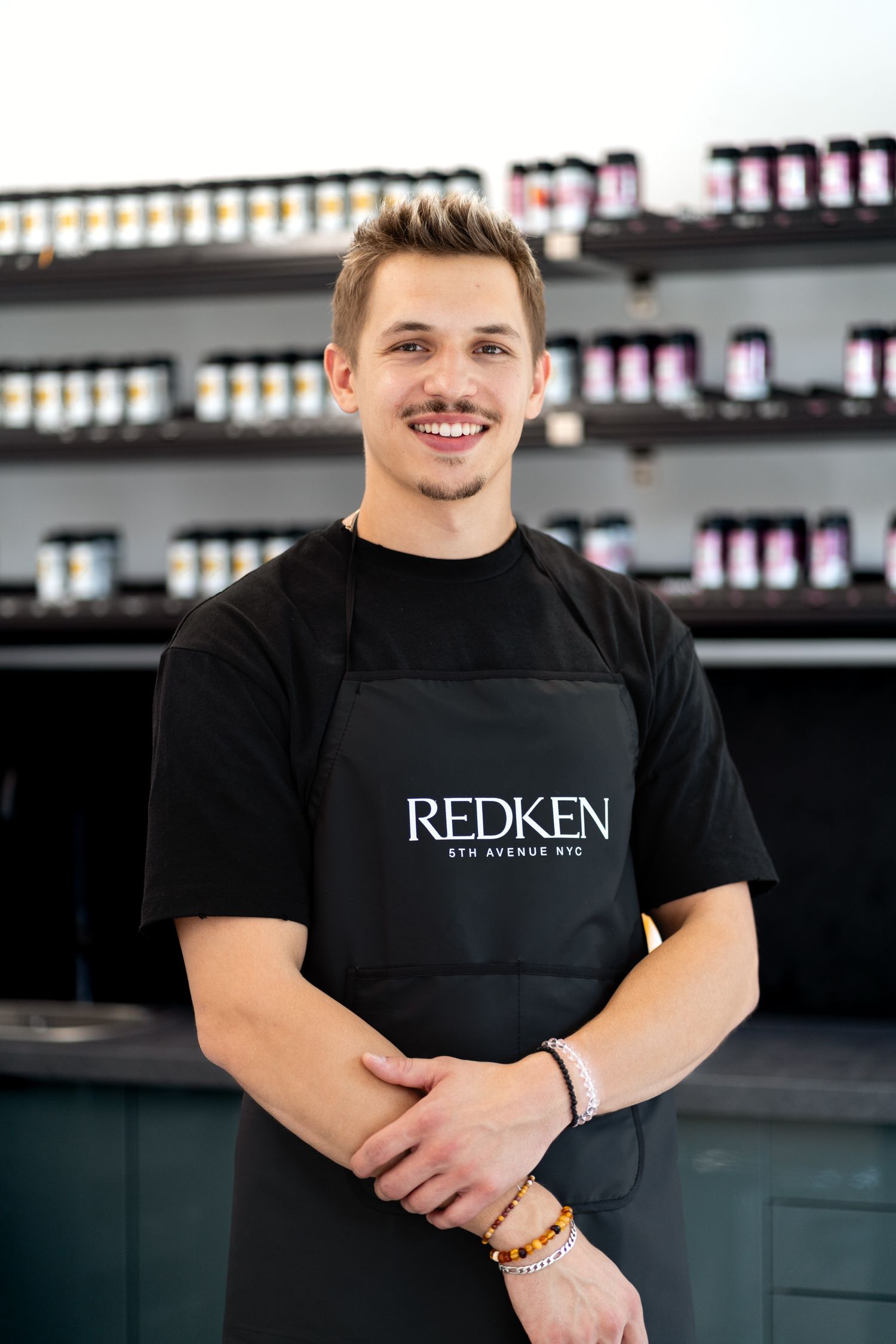Young male hairstylist smiling in black Redken 5th Avenue NYC apron in salon.