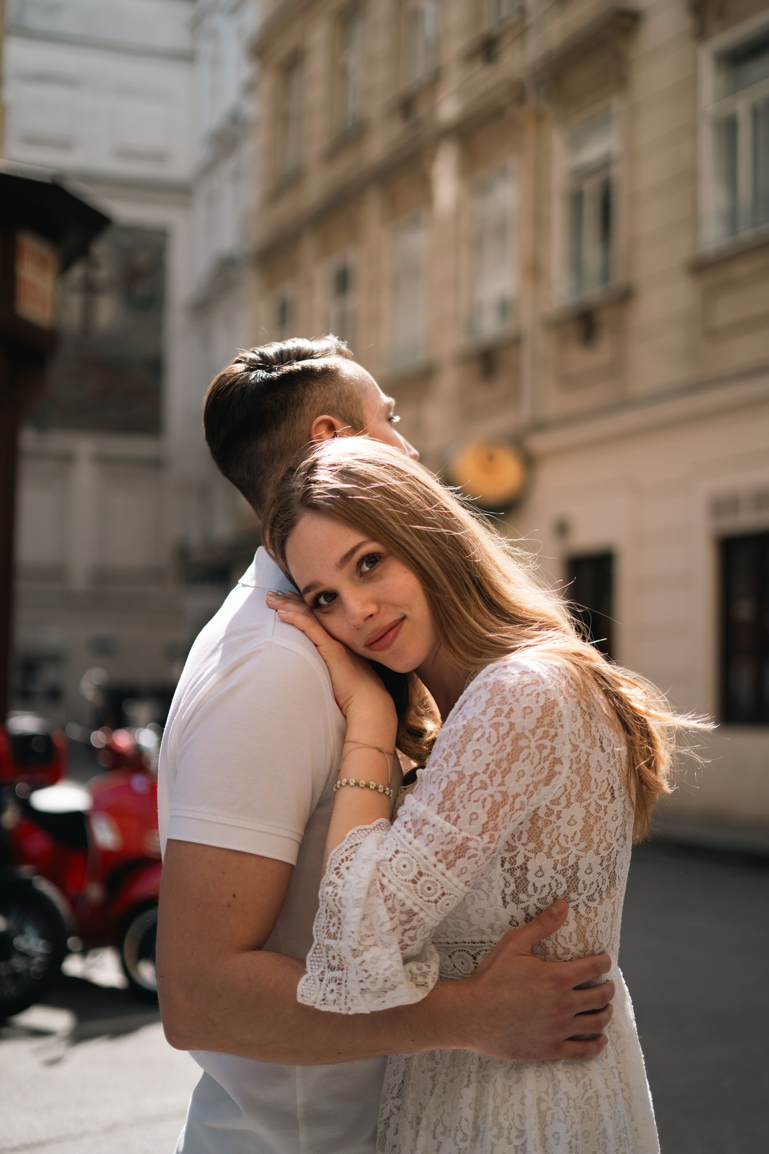 Romantic couple embracing on a sunlit European city street, woman in white lace dress.