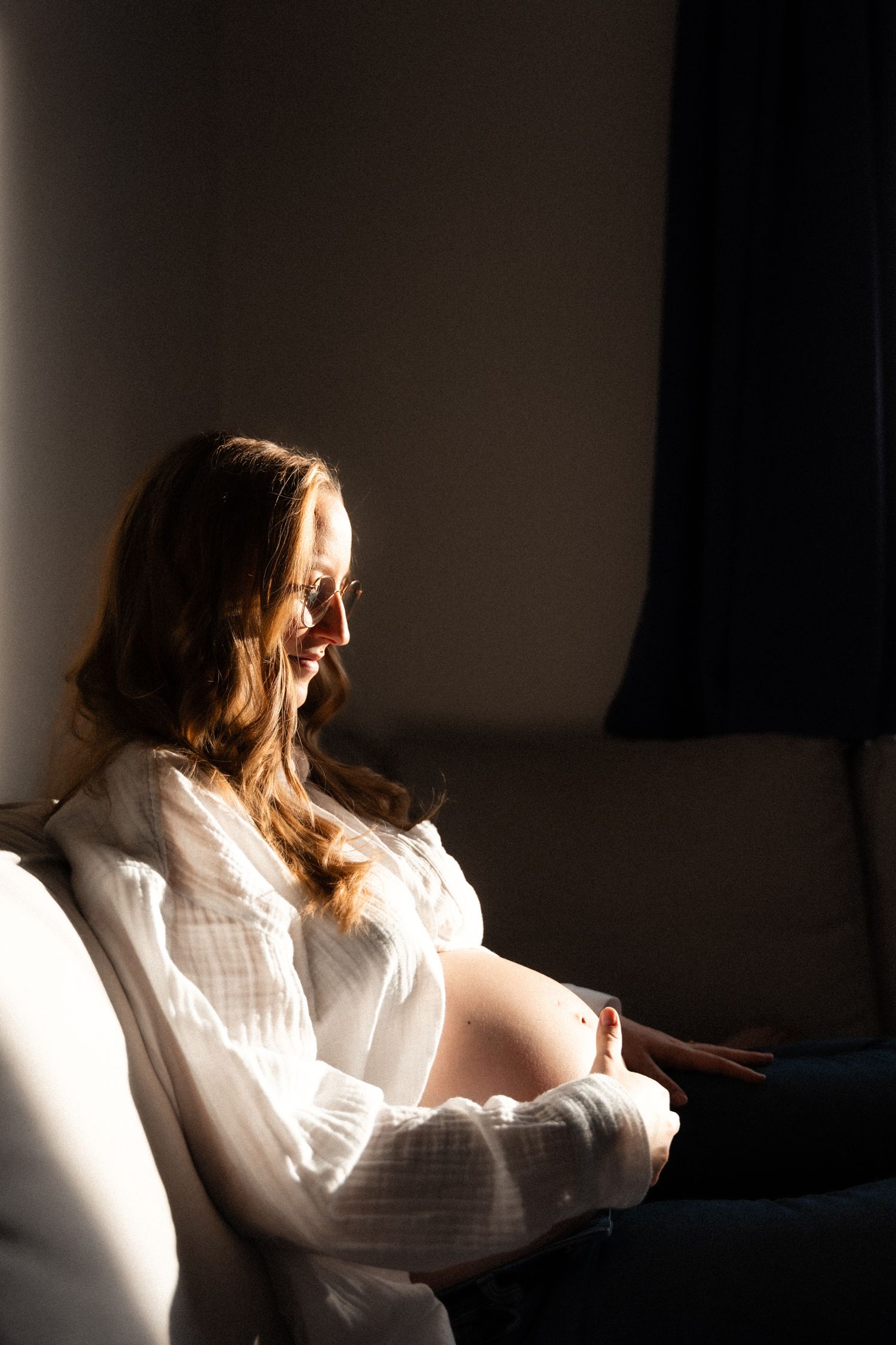 Smiling pregnant woman in white blouse sitting on sofa in warm sunlight.