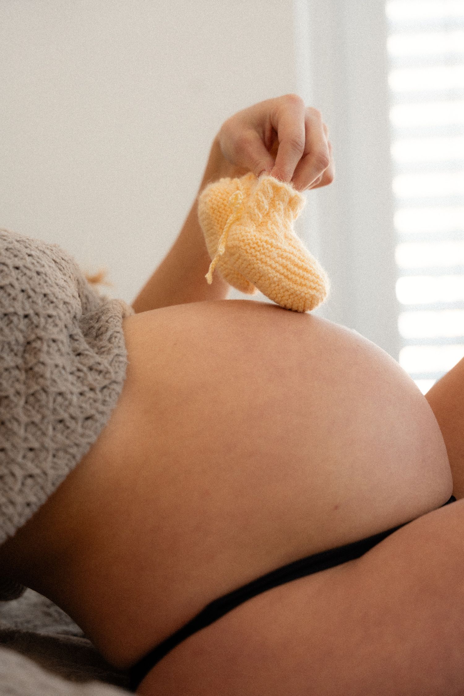 Pregnant woman lying down holding a tiny yellow knitted baby bootie above her bare belly.